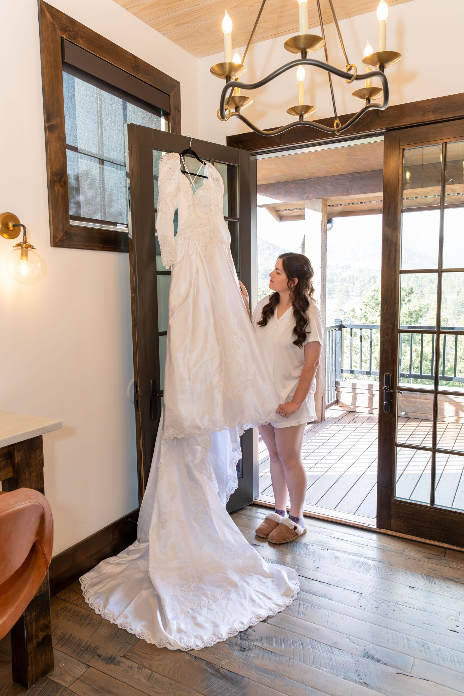 Bride in white robe admiring her wedding gown hanging in a rustic wooden room with chandelier and balcony doors.