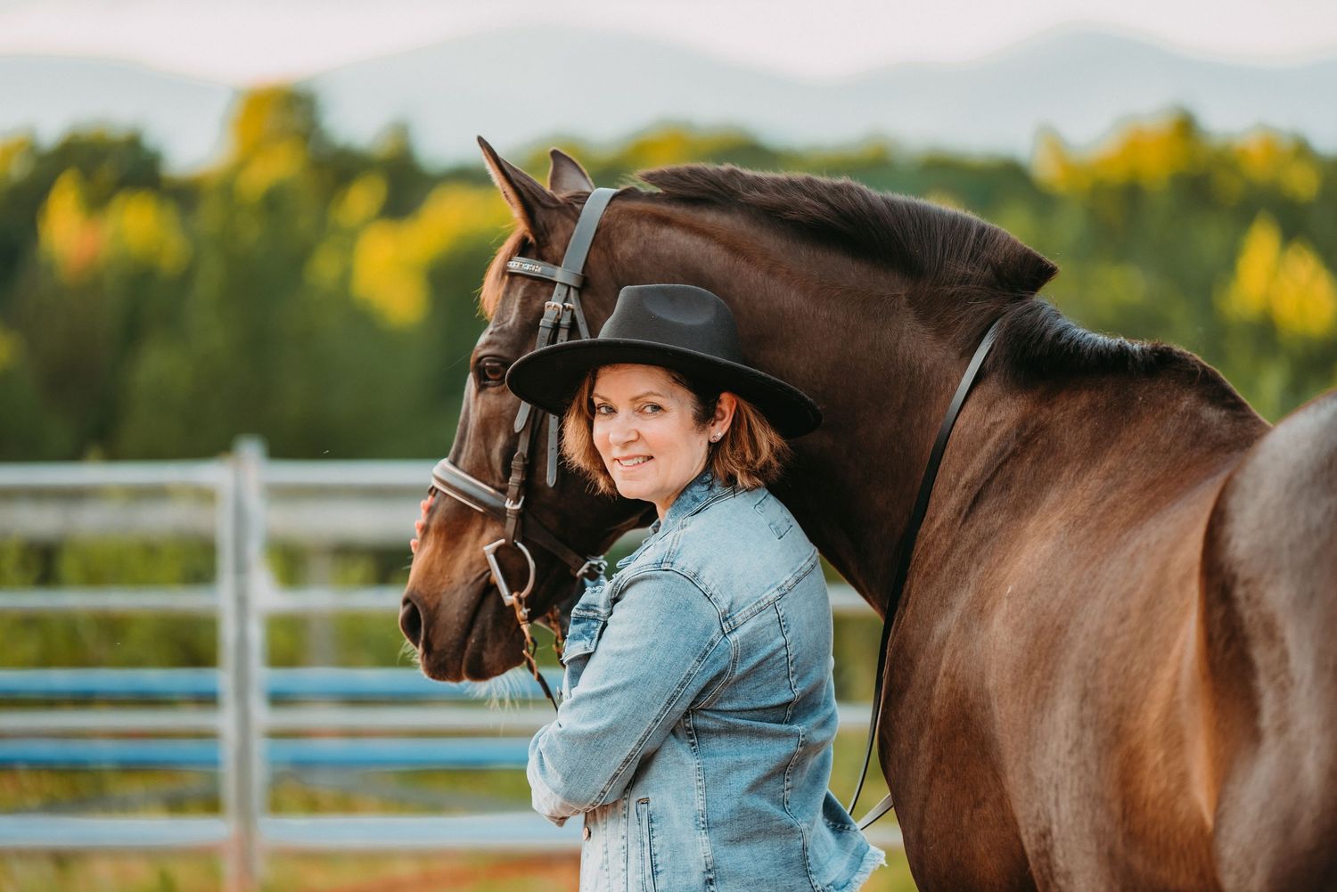Smiling woman in denim jacket and black hat leaning against a chestnut horse outdoors.