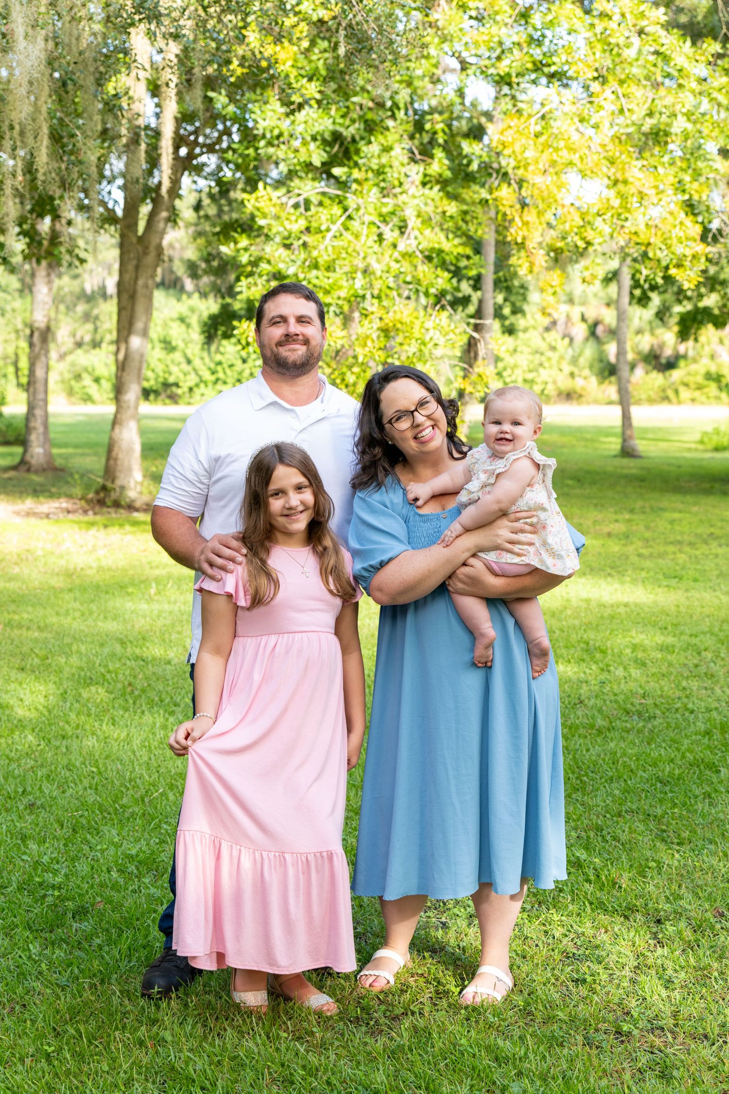 Smiling family of four posing together on a lush green lawn surrounded by trees in a park.