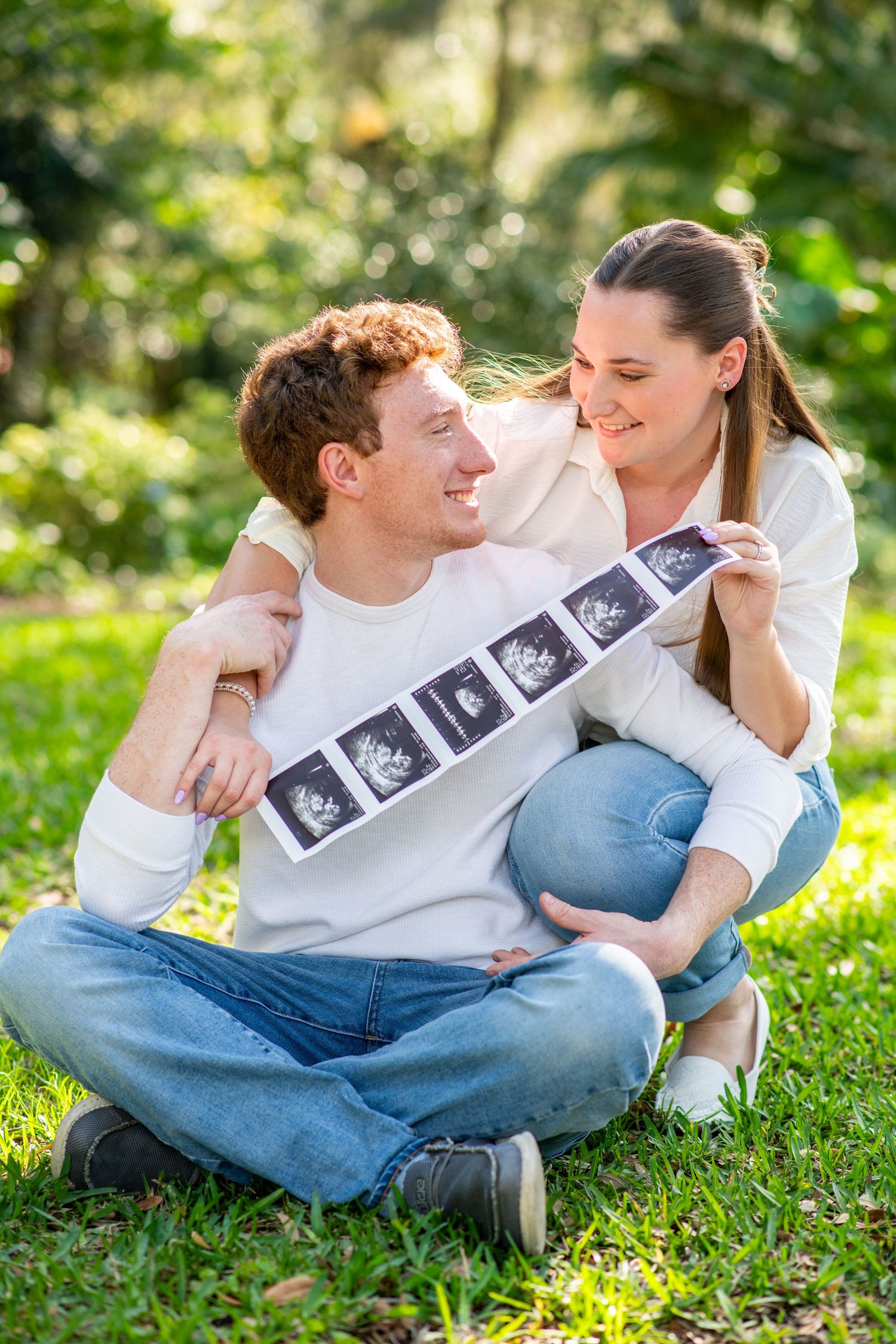 Happy couple sitting on grass in a park holding ultrasound images and smiling at each other.