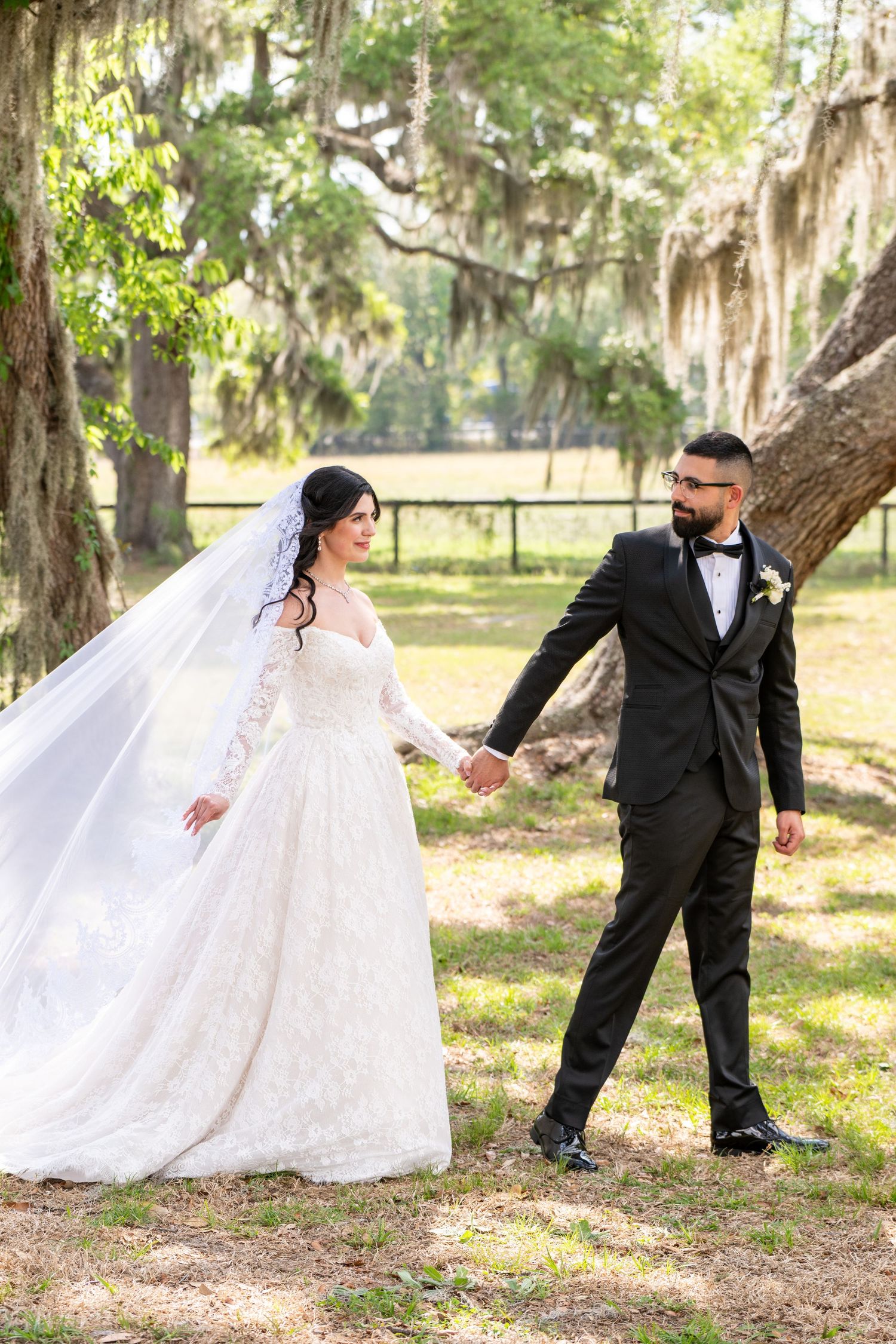 Bride and groom hold hands walking under Spanish moss-draped trees in a sunlit outdoor wedding photo.
