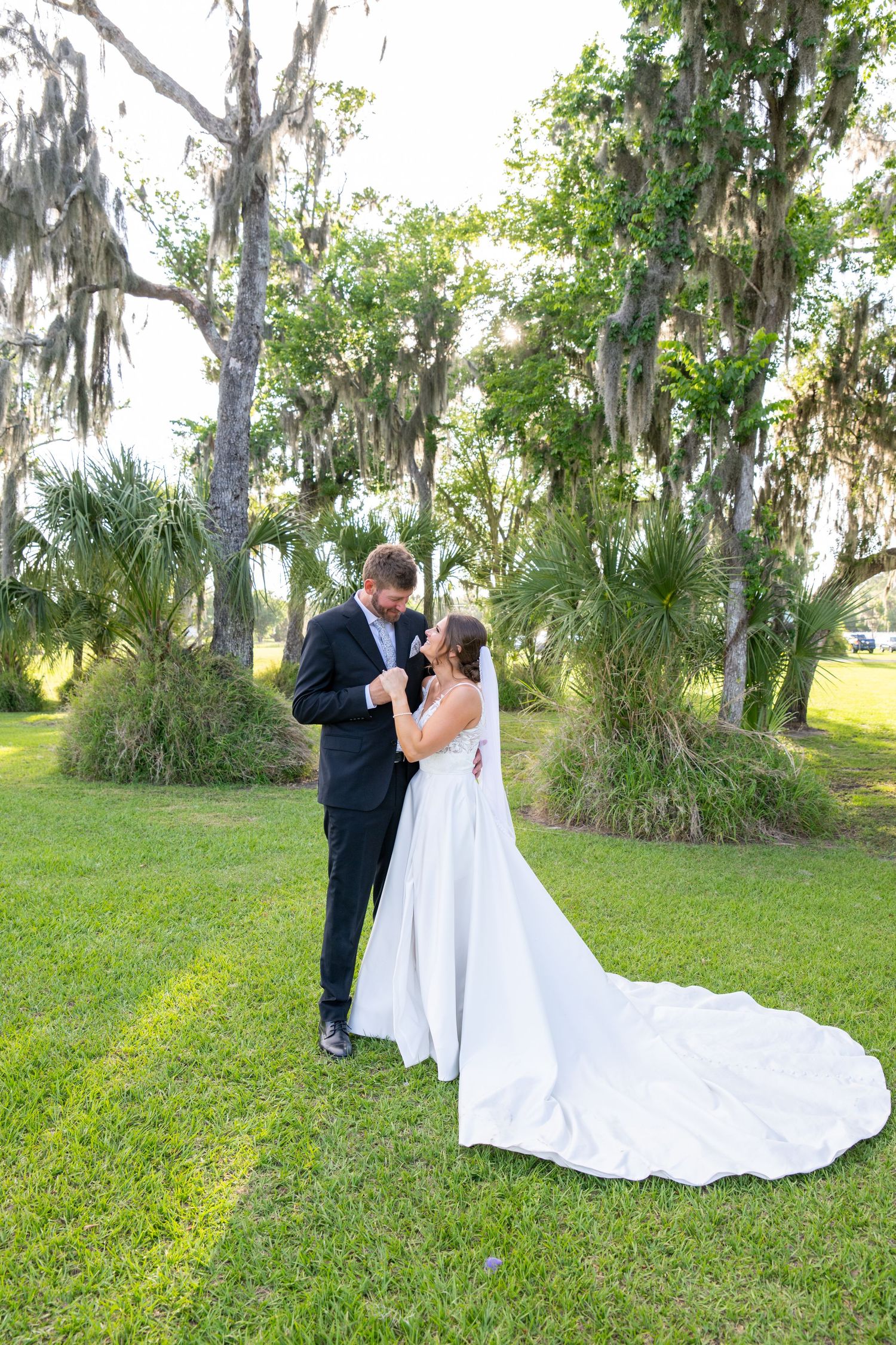 Bride and groom share a romantic moment on lush green lawn surrounded by moss-draped oak trees in elegant wedding attire.
