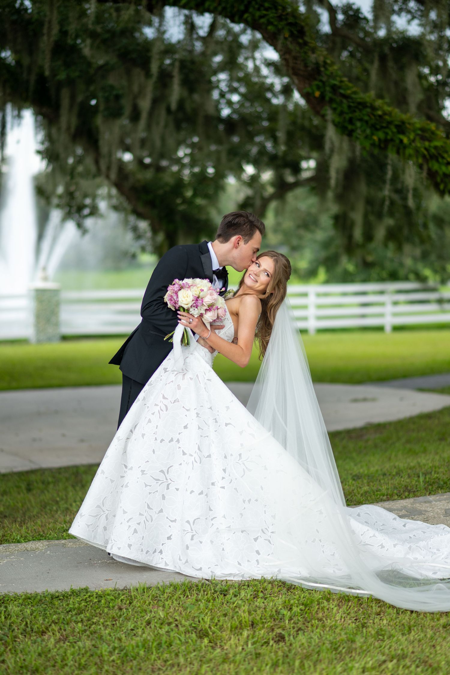 Groom kisses bride in elegant white ballgown holding pink bouquet outdoors near white fence and moss-draped oak tree.