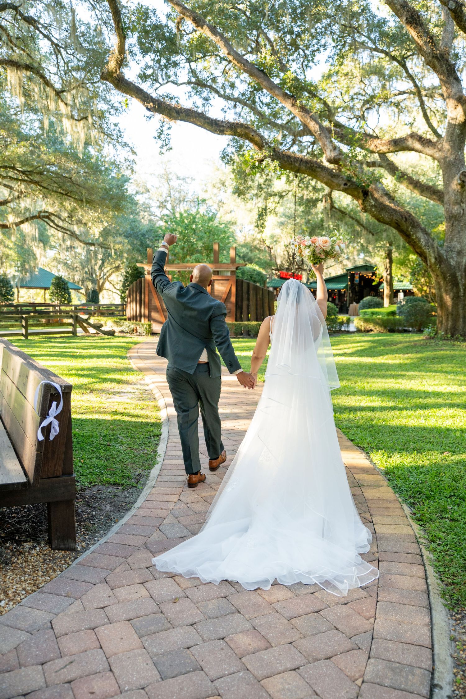 Bride and groom walking hand-in-hand down a brick path under oak trees, groom raising fist in celebration.