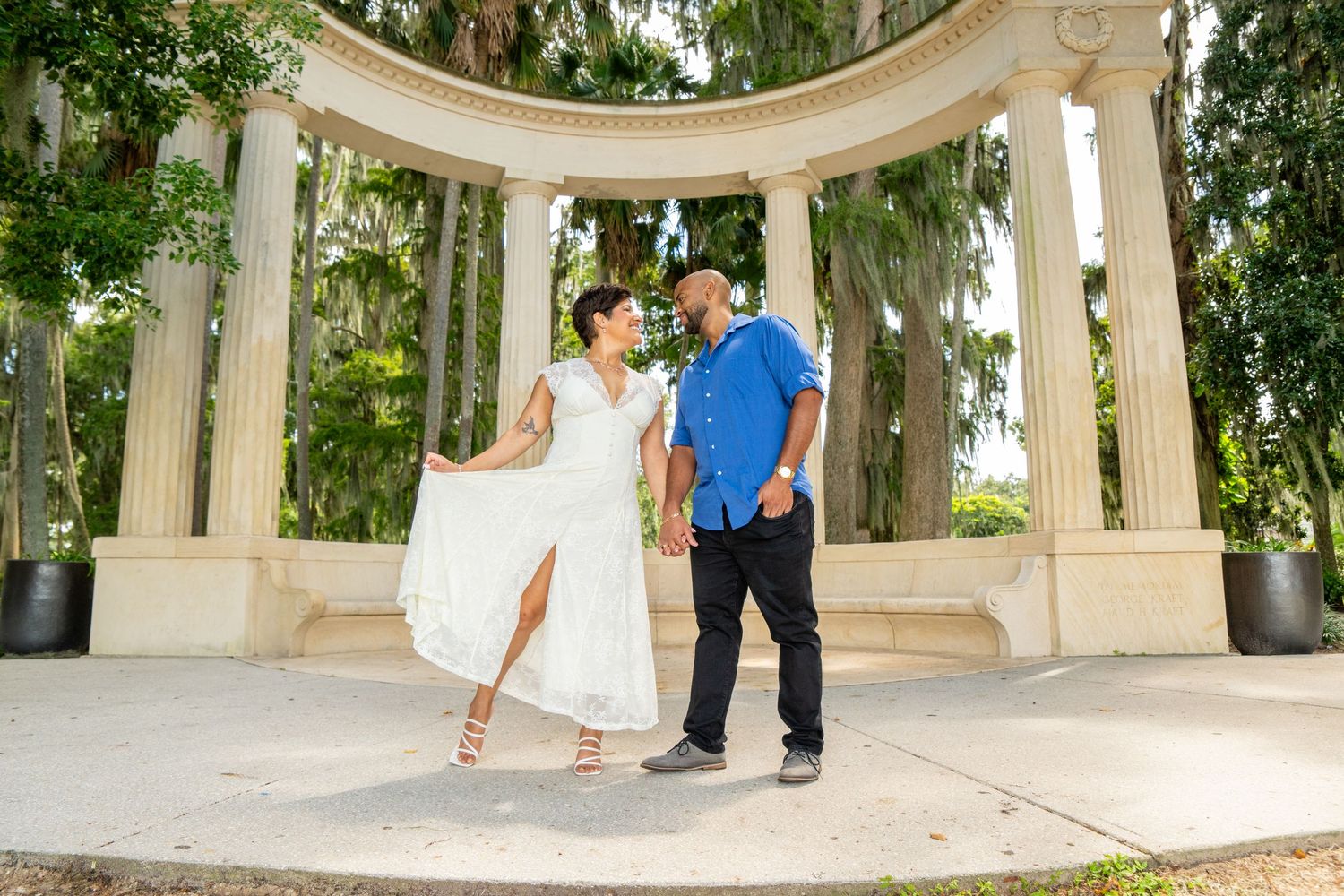 Couple dancing and holding hands under elegant white columns surrounded by lush green trees outdoors.
