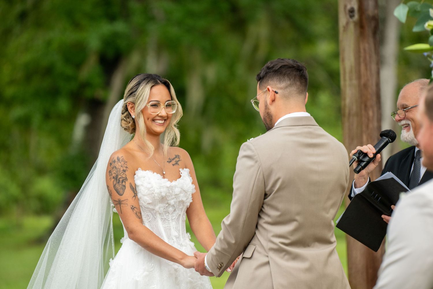 Smiling bride in white strapless gown and veil holds hands with groom in tan suit during outdoor wedding ceremony.