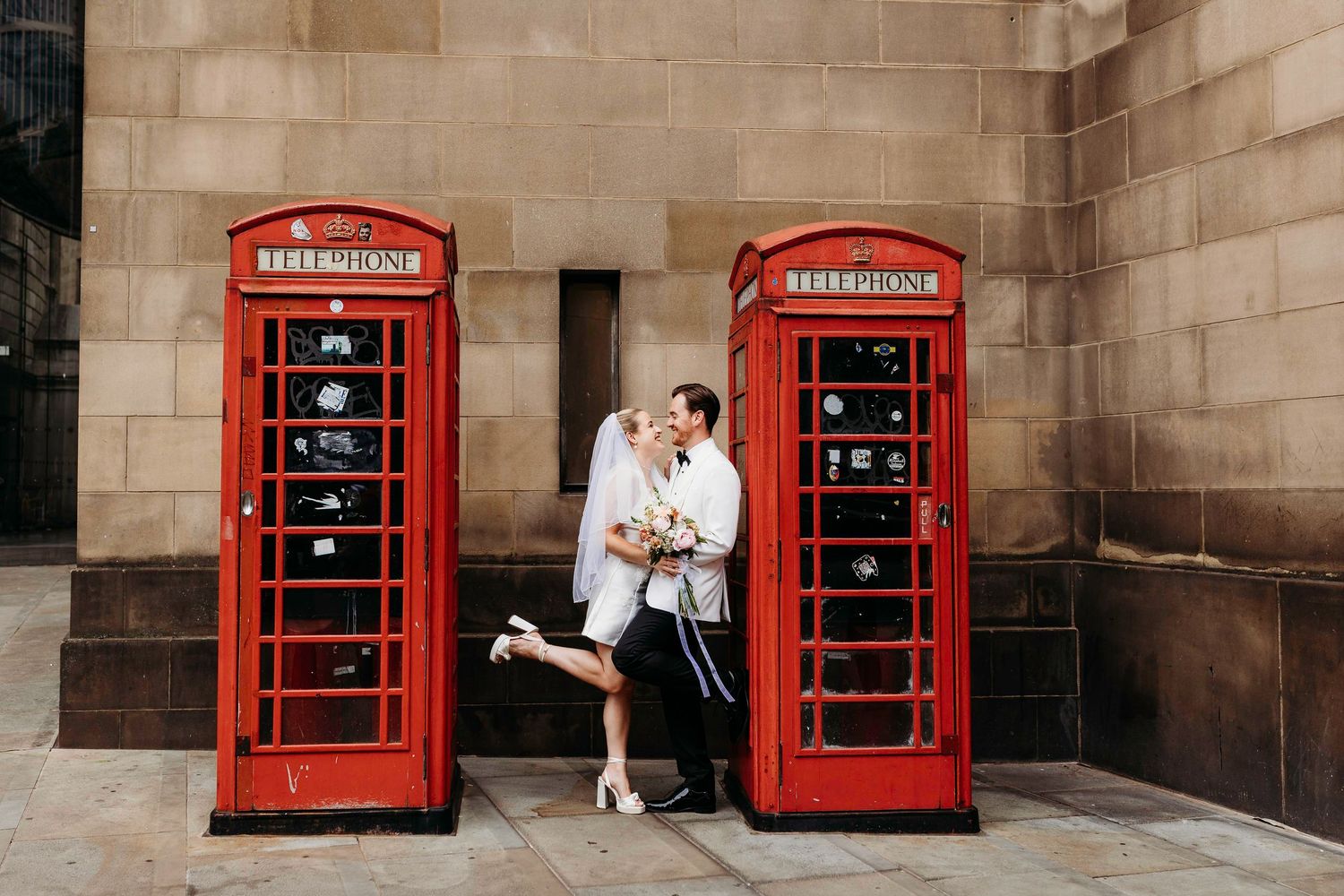 Wedding Couple with classic red British phone booths against stone building backdrop in Manchester City Centre Elopement.