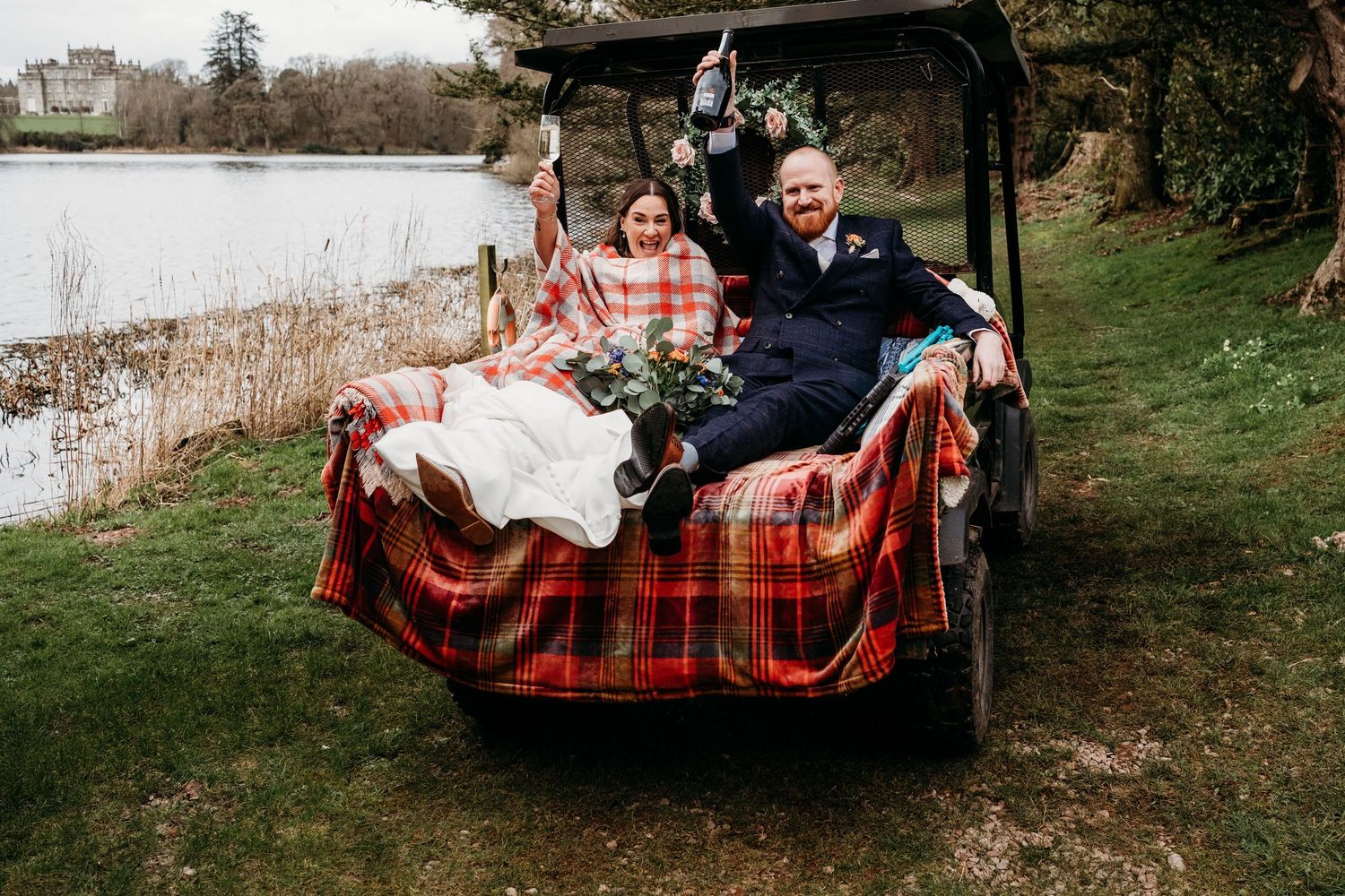 Bride and groom riding a tartan-draped golf cart by a lake, celebrating their Scottish wedding outdoors.