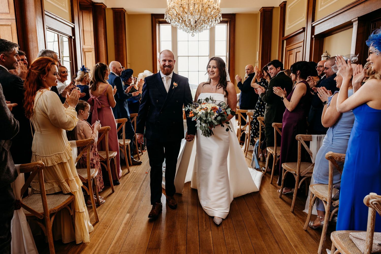 Bride and groom walk down the aisle at elegant indoor wedding ceremony with chandelier and applauding guests.