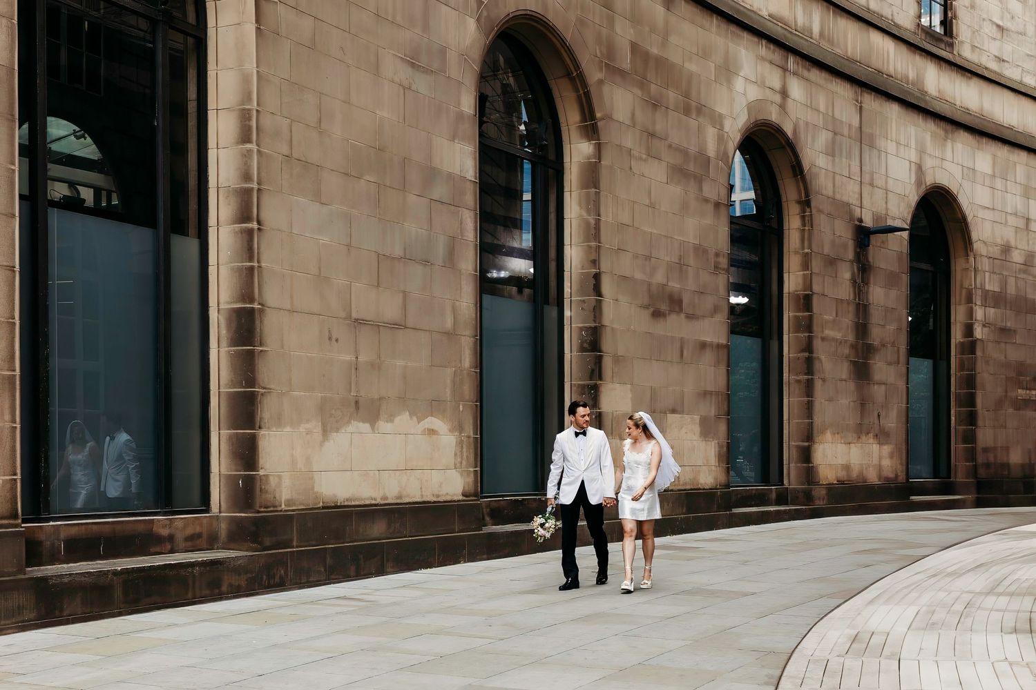 Bride and Groom walk along a curved stone building exterior with arched windows and columns in Manchester City Centre Wedding Celebrations.