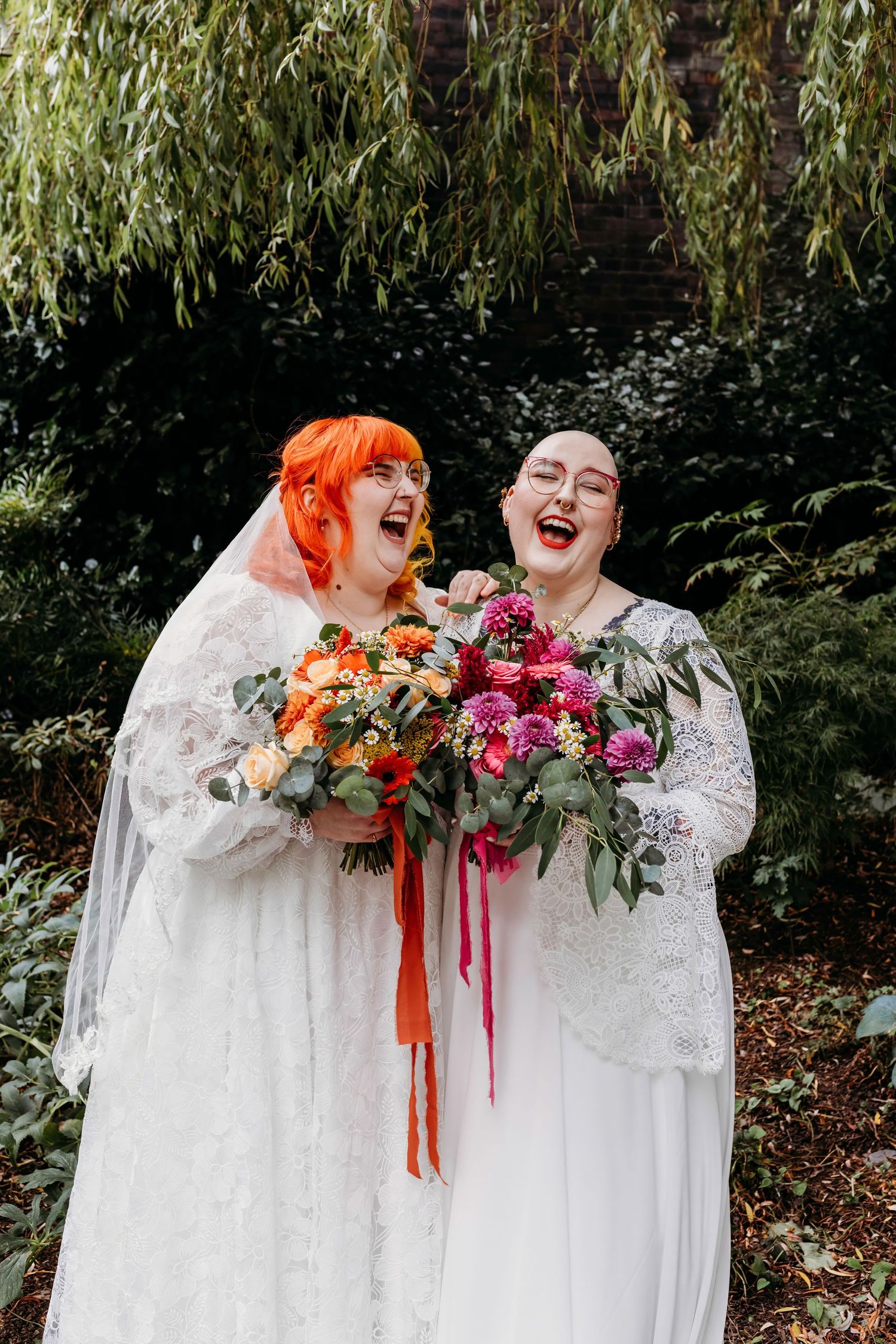 Two brides in white dresses holding vibrant floral bouquets under a willow tree at Spode China Hall Gardens, Stoke.