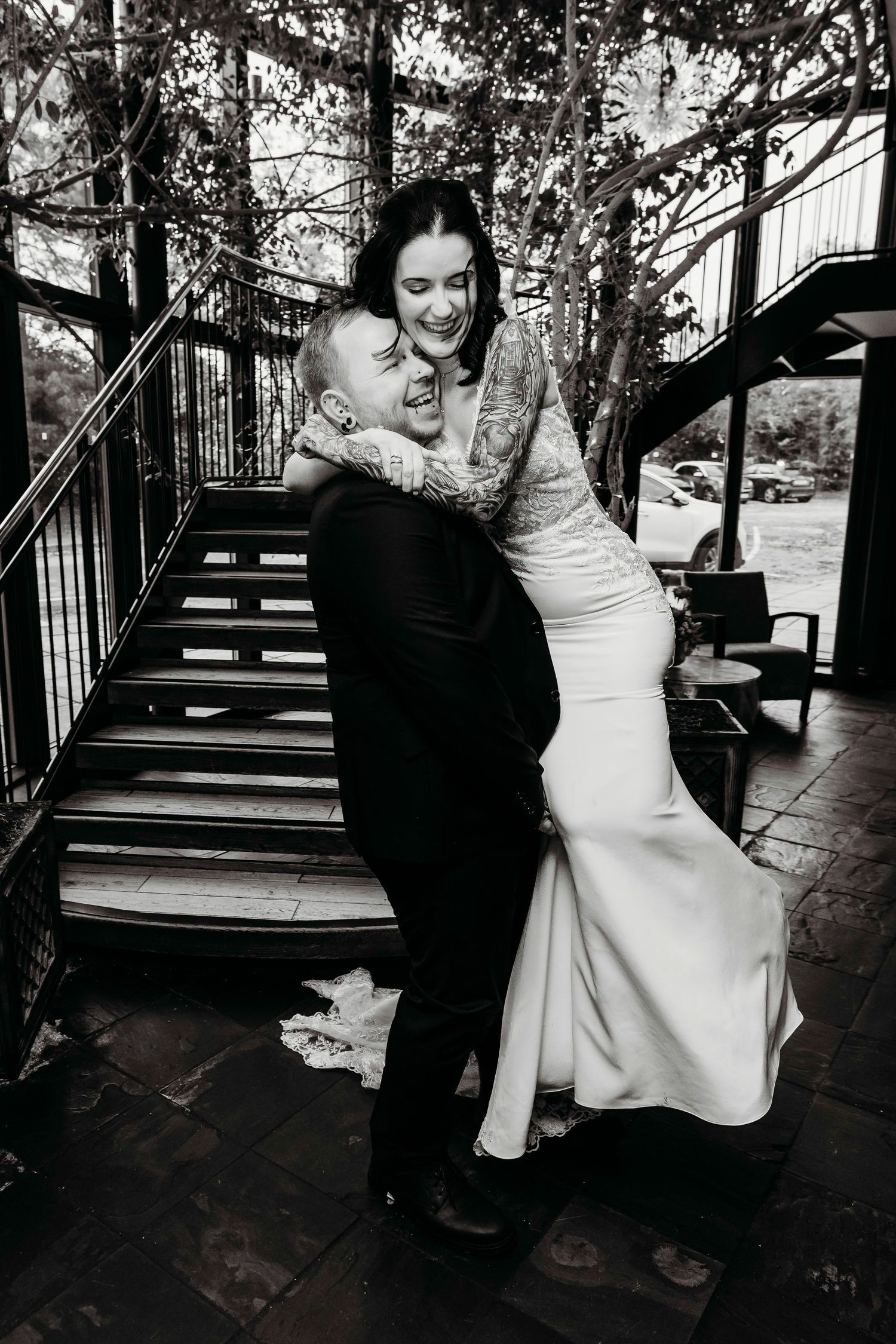 Black and white photo of a couple embracing on outdoor wooden stairs, with the bride wearing an elegant fitted white gown.