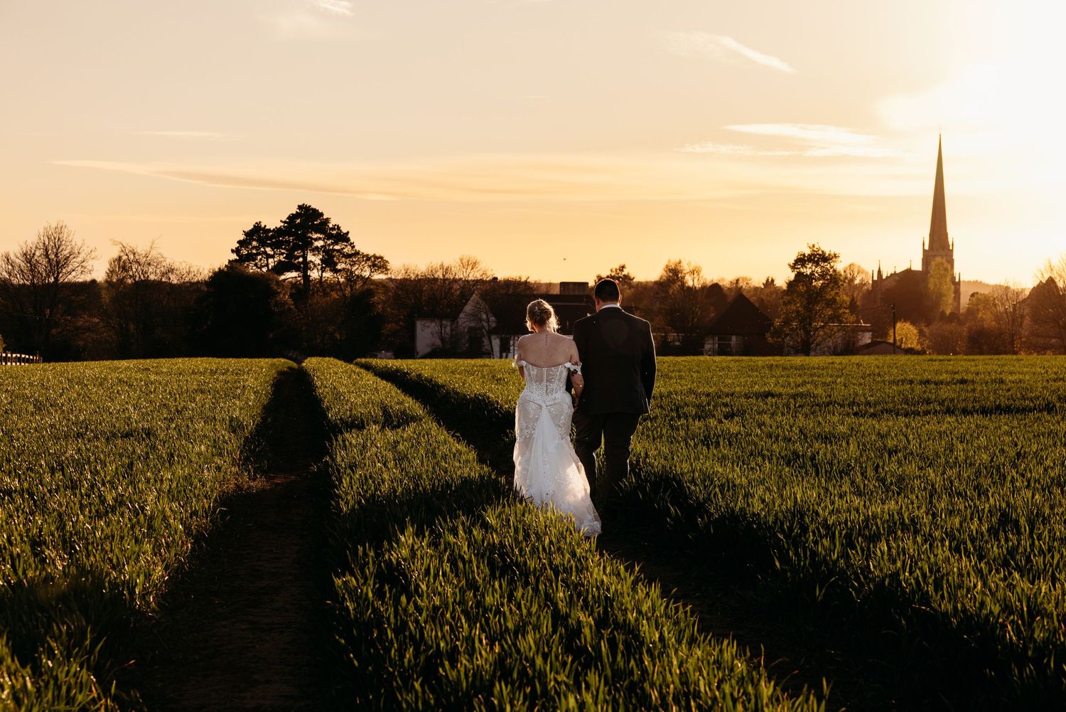 Bride and groom walking through green fields at golden sunset with church spire in background.