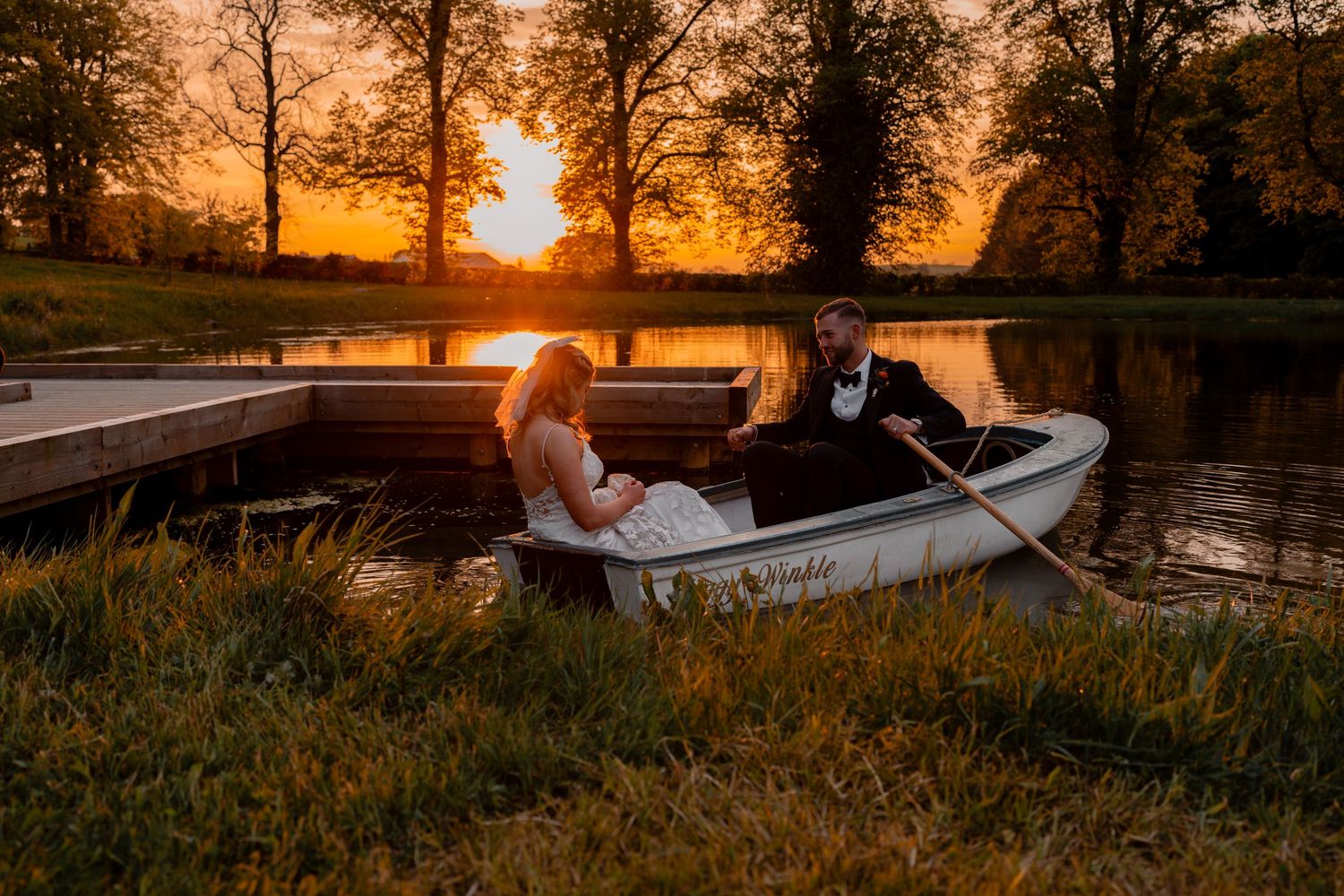 Newlyweds take a small boat on calm water at sunset in the Lake District at Holesfoot wedding venue silhouetted trees in the background.