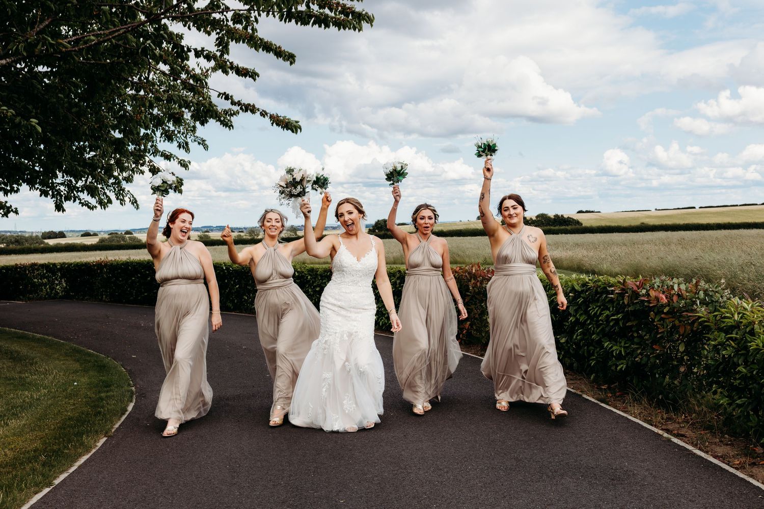 Bride and Bridesmaids in beige dresses pose together on a countryside path under blue skies at Burntwood Court Wedding Venue, Yorkshire.