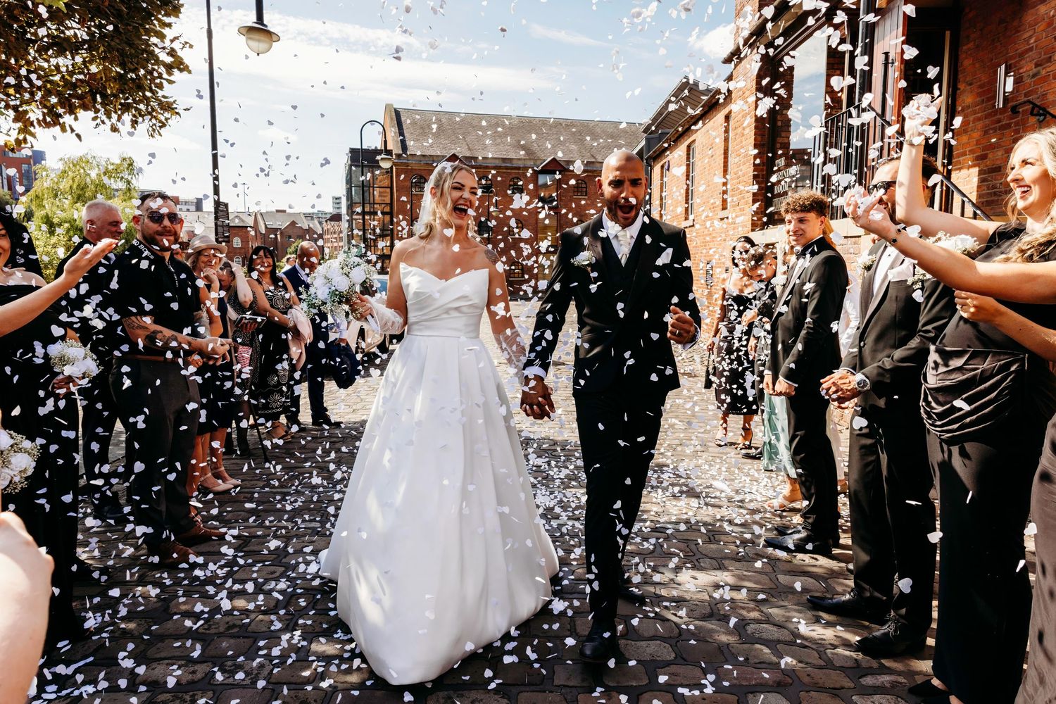 Wedding guests throw white confetti over the newlyweds during their joyful outdoor celebration exit at the Castlefield Rooms in Manchester.
