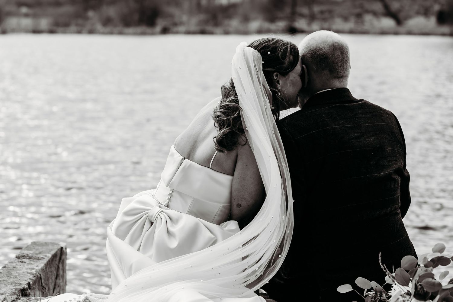 Black and white photo of a bride and groom sitting together by a lake, sharing an intimate moment.