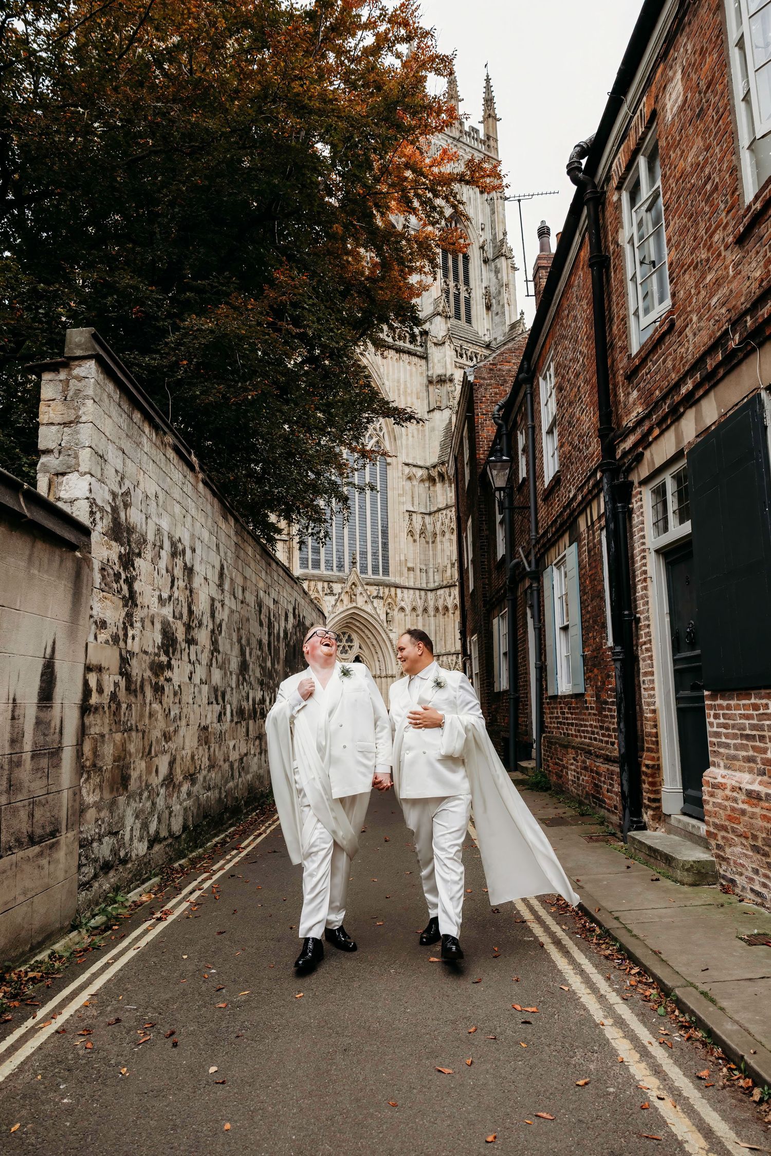 Two Grooms in white outfits walk along a historic York stone-walled alley with the iconic York Minster visible in the background.