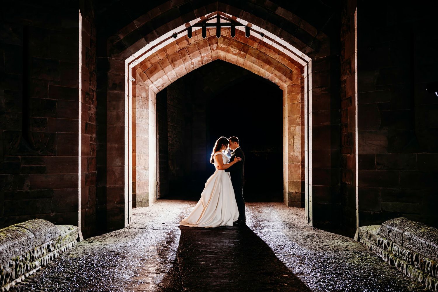 A romantic silhouette of newlyweds embracing in a stone archway of Peckforton Castle with dramatic backlighting creating a glowing effect.
