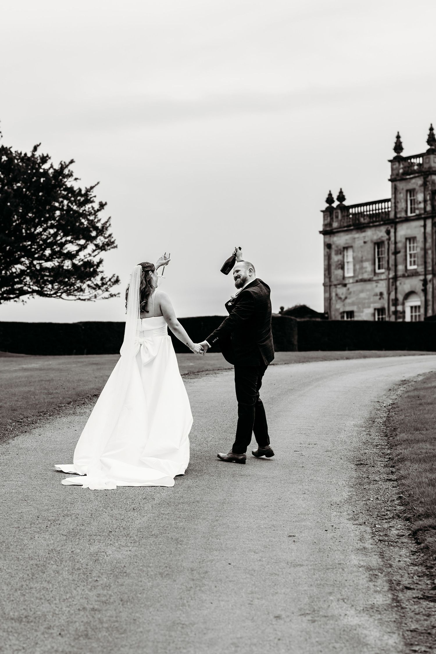 Black and white photo of bride and groom holding hands, dancing on a path outside a grand historic estate.
