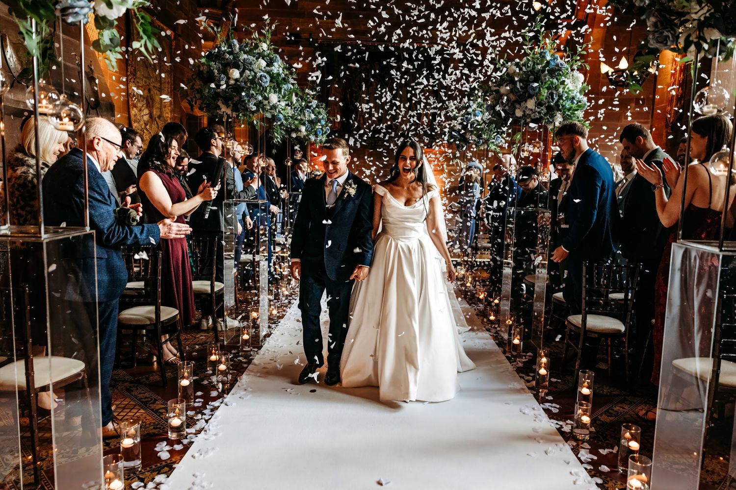 Wedding couple walking down aisle at Peckforton Castle, Cheshire, while guests throw white confetti in candlelit venue.