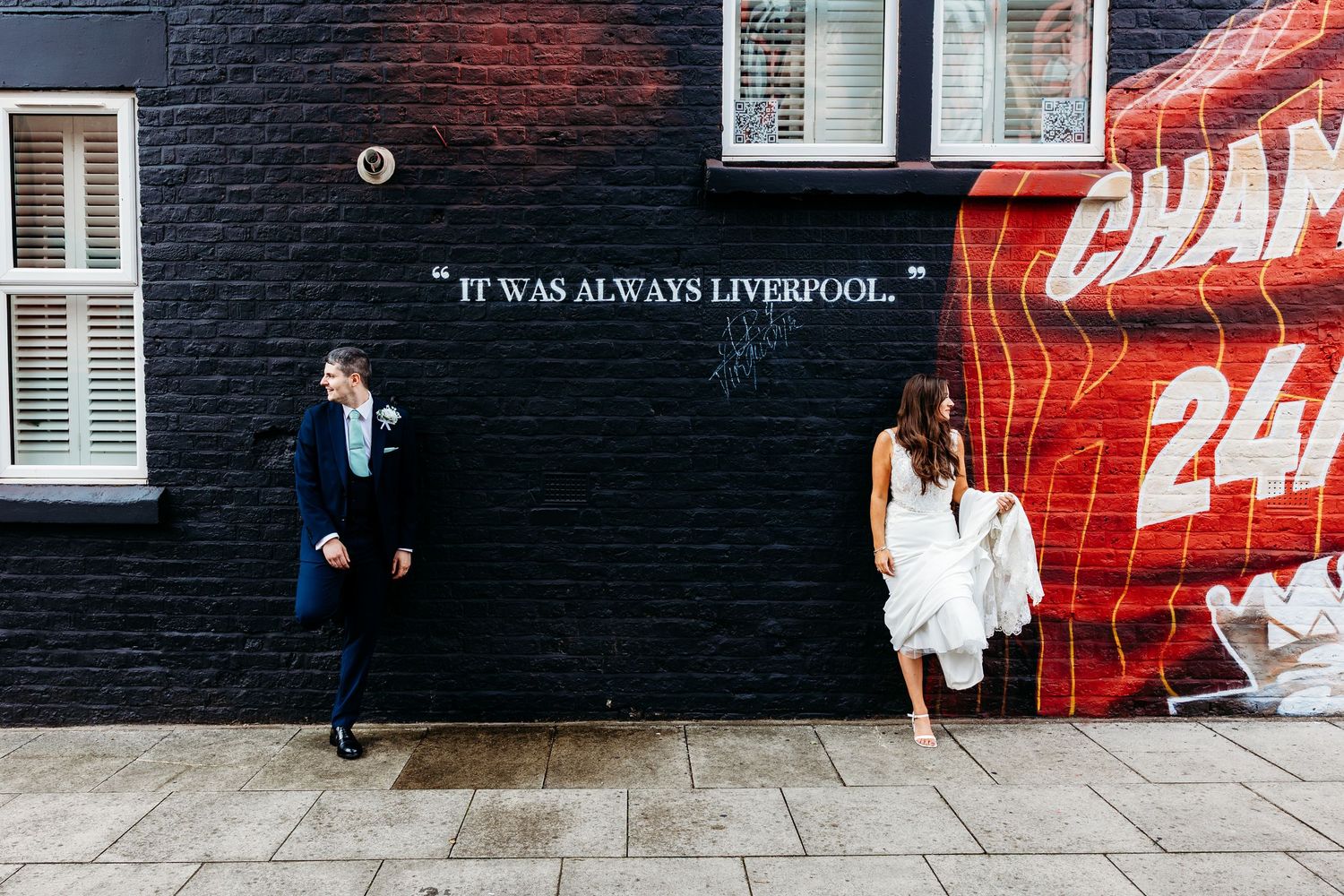 Bride and Groom portraits with Urban street art featuring Liverpool text on black brick wall with red accents near Anfield, Liverpool.