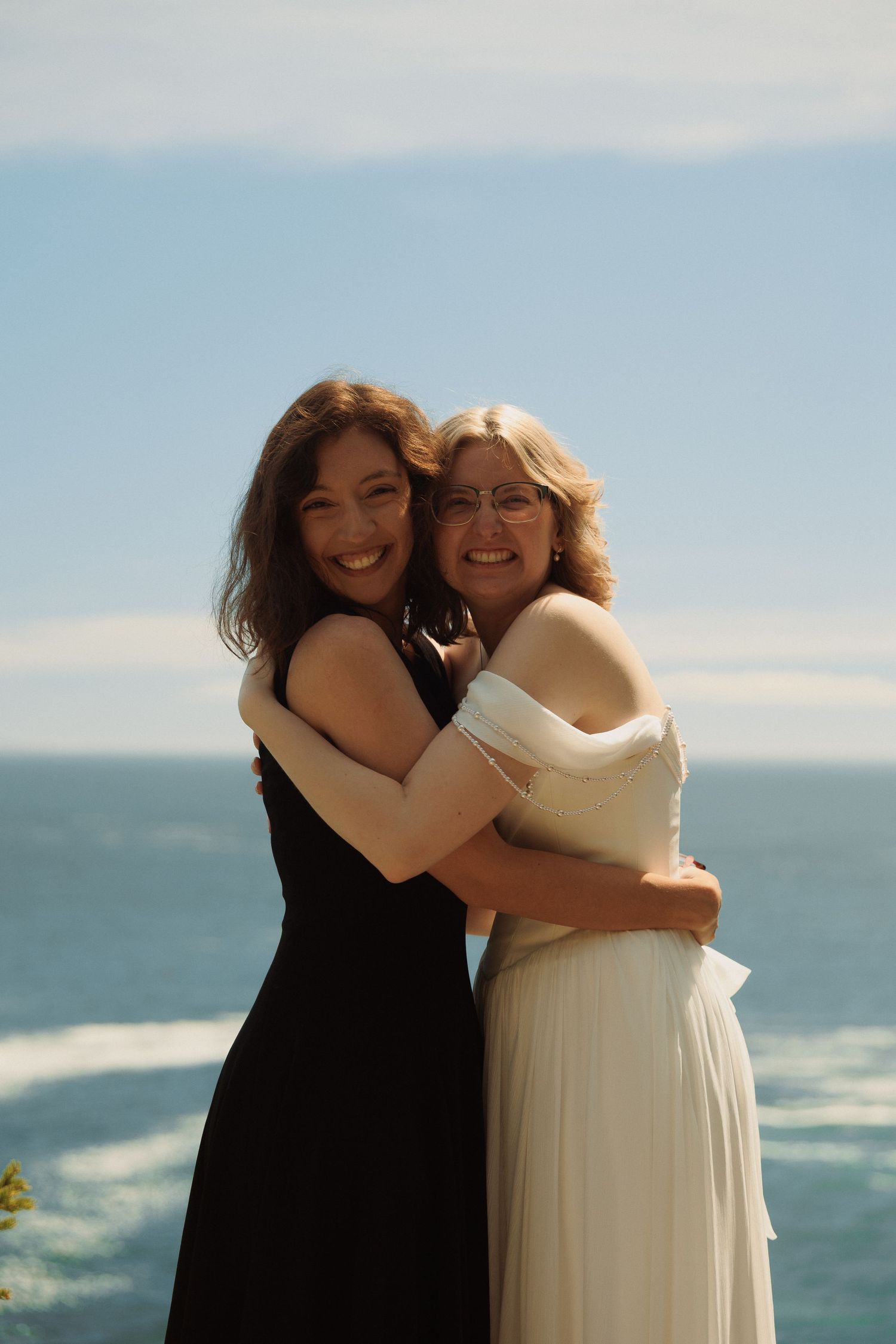 Two women embrace and smile in formal attire with ocean backdrop at a wedding.