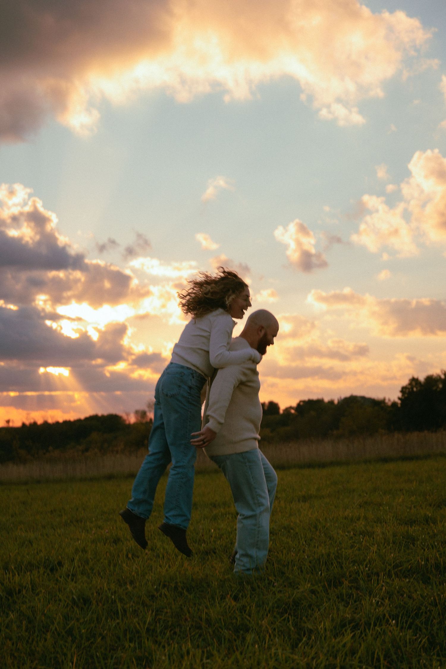 Two people playfully interact in a field during a dramatic sunset with orange clouds.