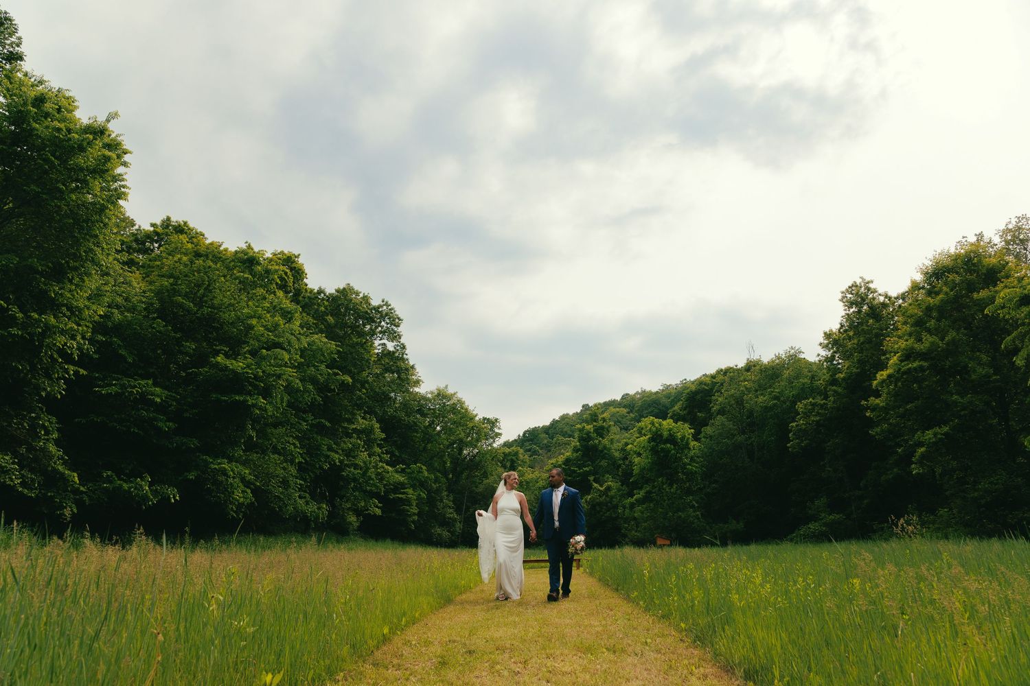 A couple walks down a path through a lush green meadow surrounded by trees on a cloudy summer day.