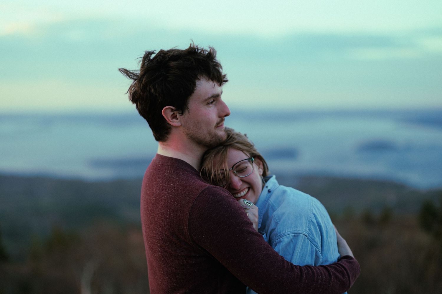 A young couple embraces warmly at sunset, smiling together against a scenic landscape backdrop.