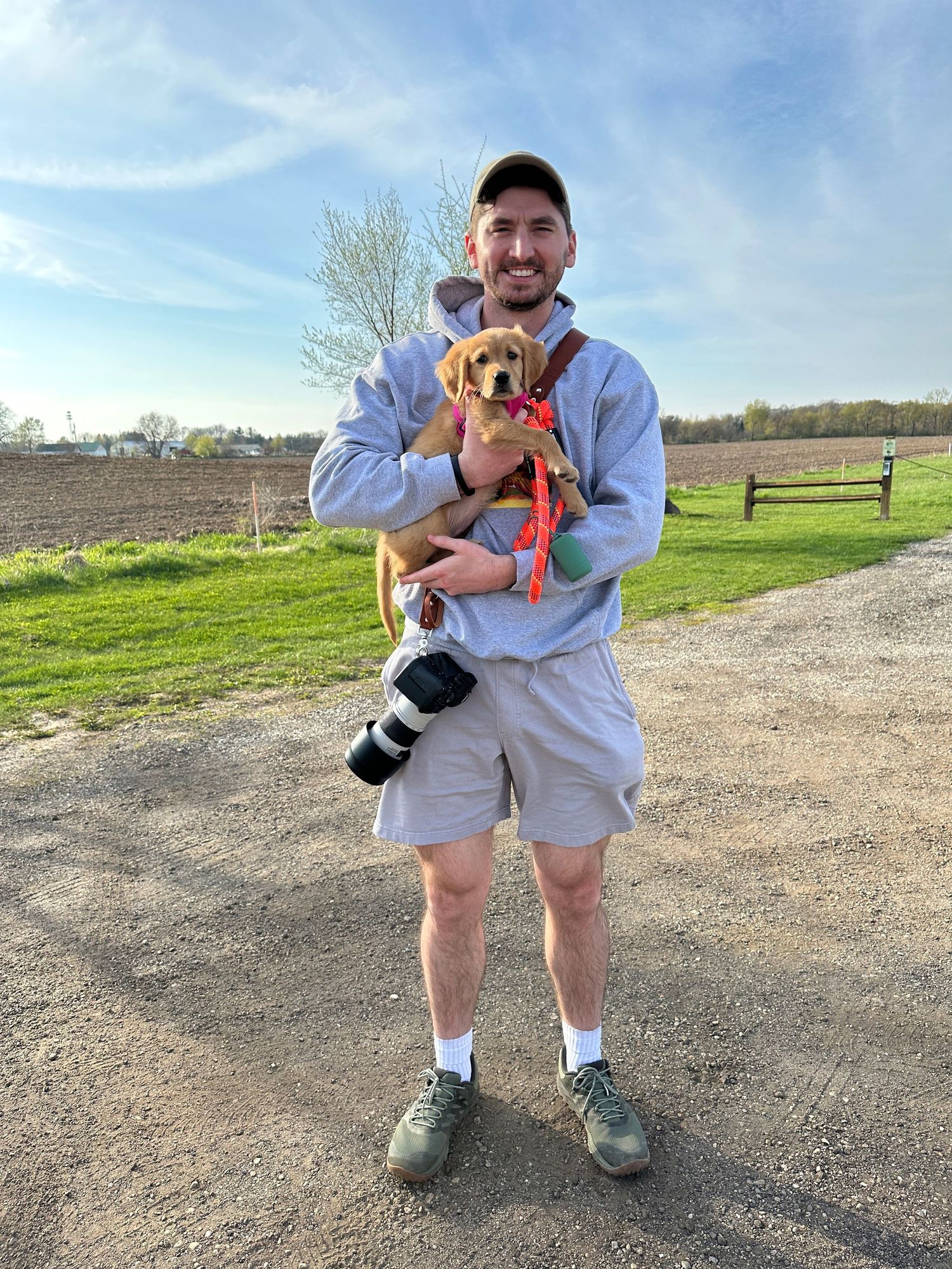 A person in casual attire holding a small dog while standing on a dirt path with green fields and trees in the background.