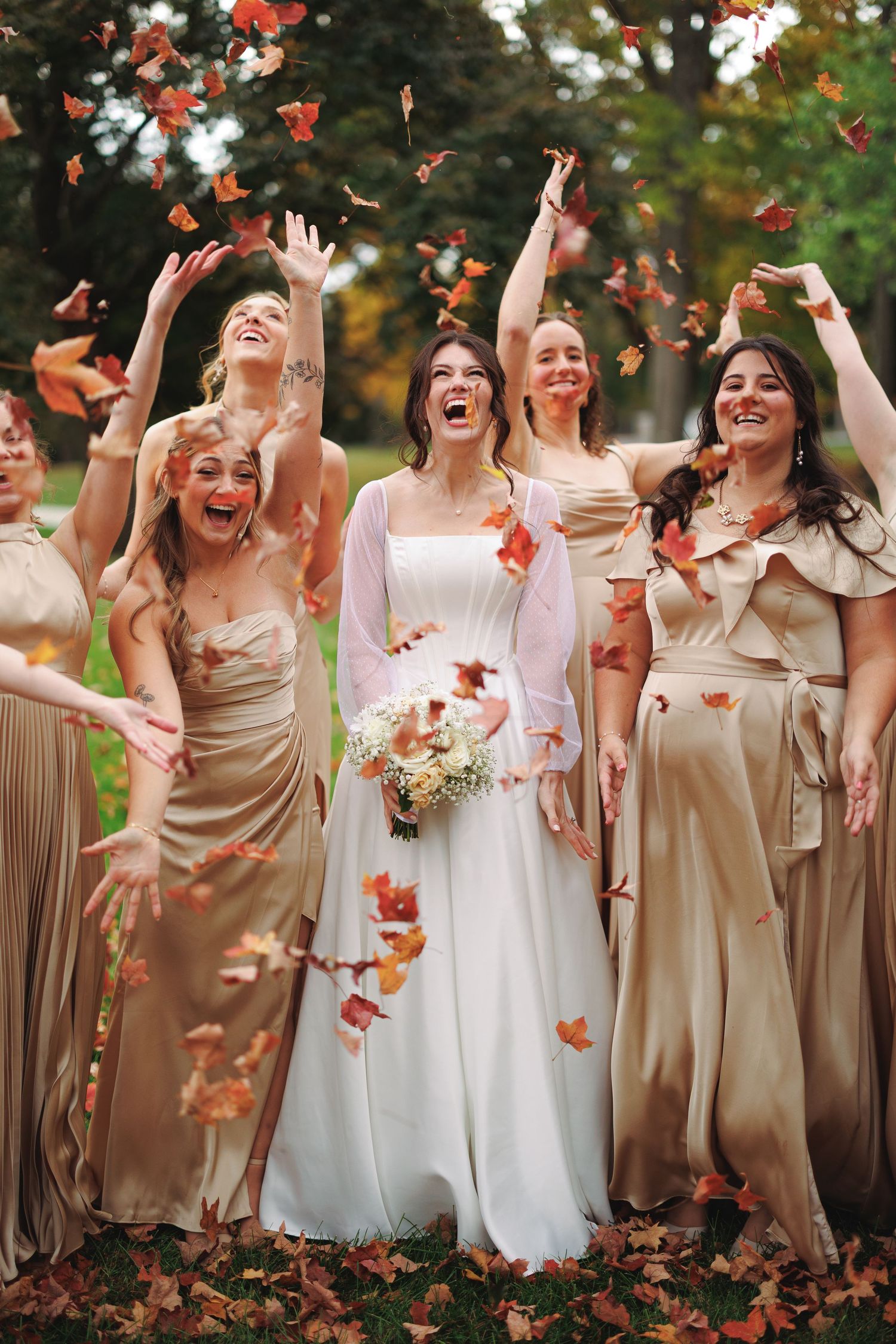 Bridesmaids in champagne dresses tossing fall leaves in joyful celebration.