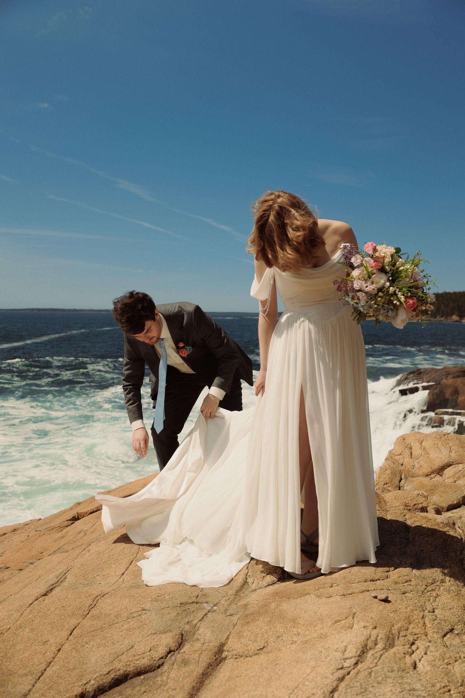 Bride and groom pose on rocky coastal cliffs, groom adjusting wedding dress train with ocean waves crashing behind them.
