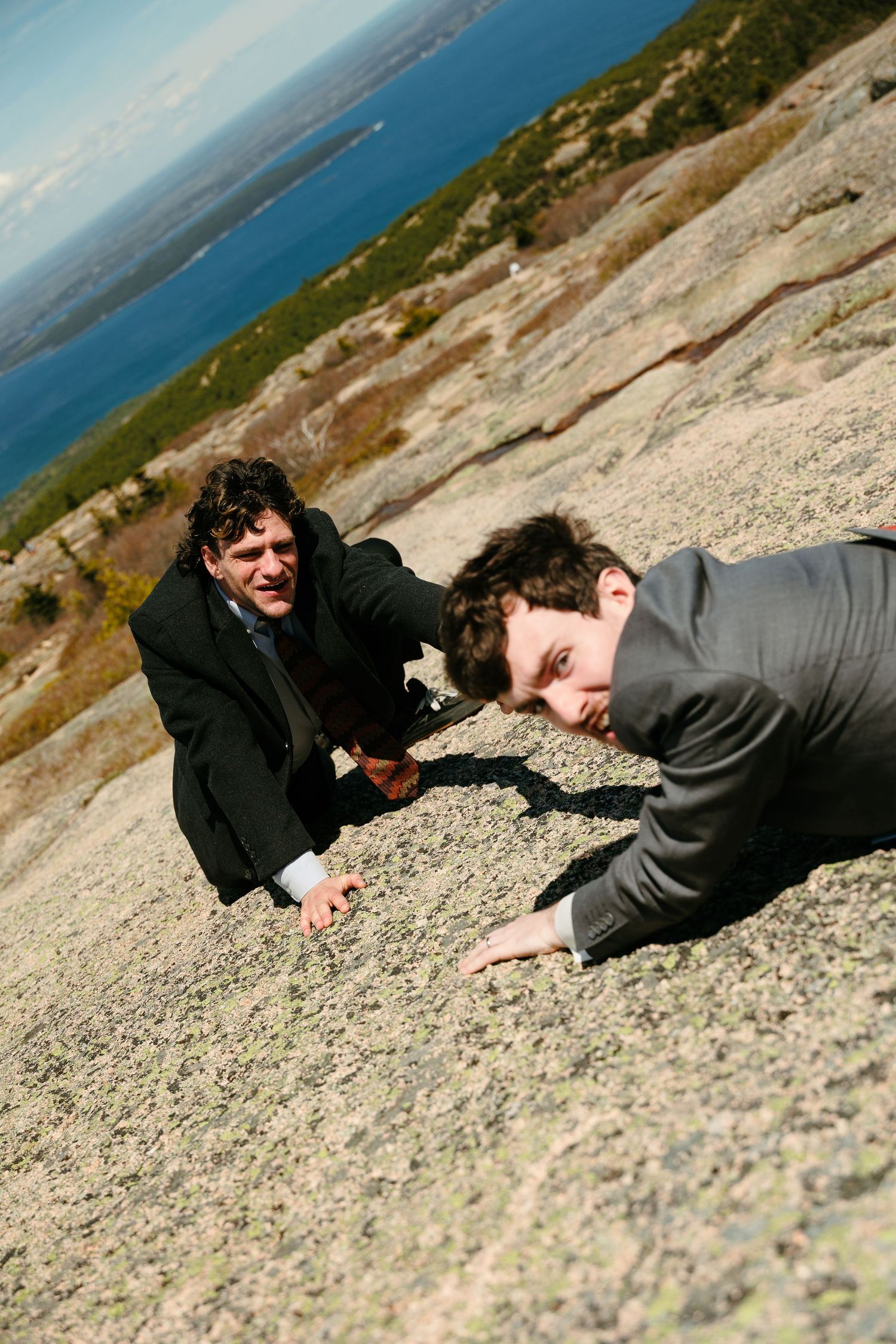 Two grooms in suits playfully crawl up a granite mountain slope at their wedding.