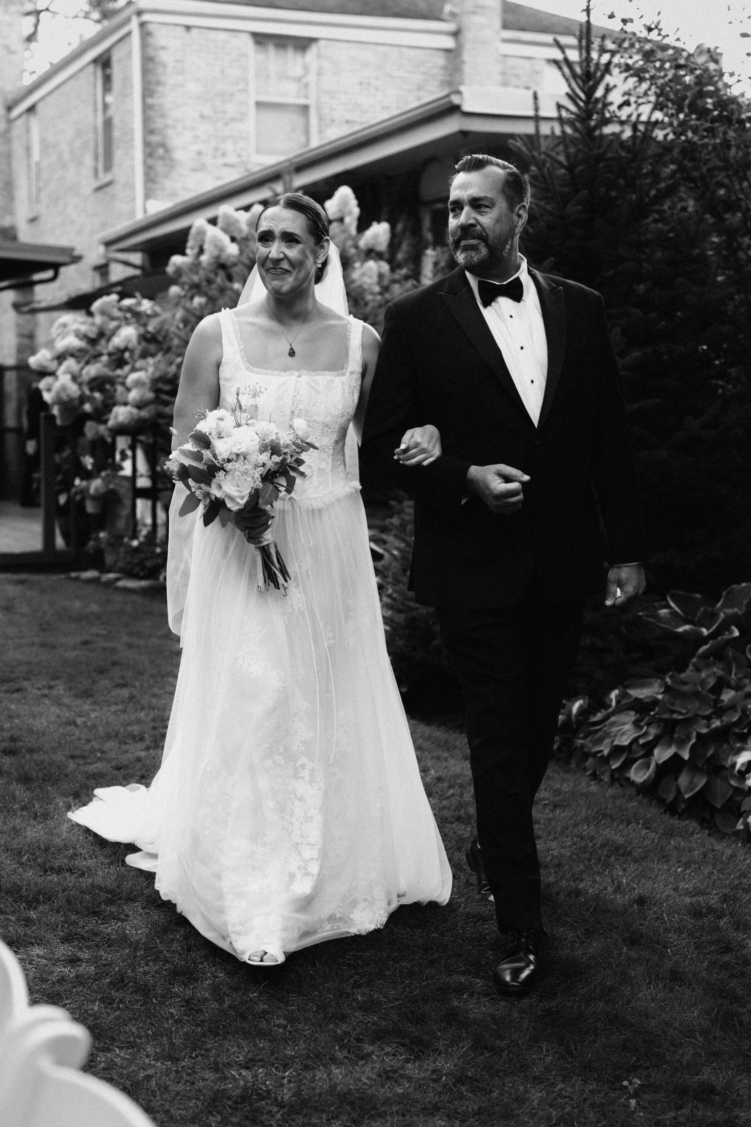 Wedding couple walking through garden with classic brick building in background.