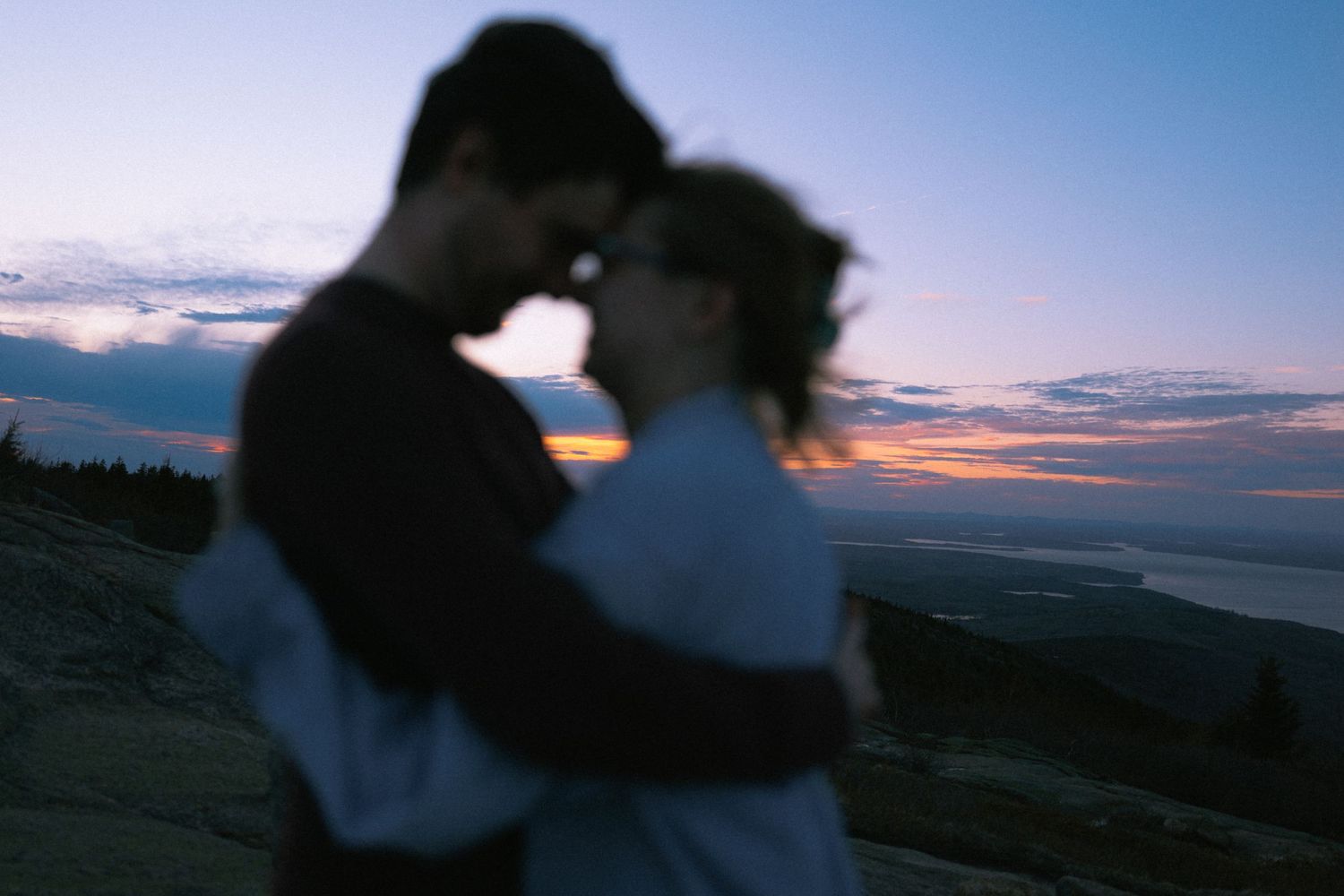 A romantic couple embraces at sunset on a hilltop, silhouetted against a colorful dusk sky with scenic views.
