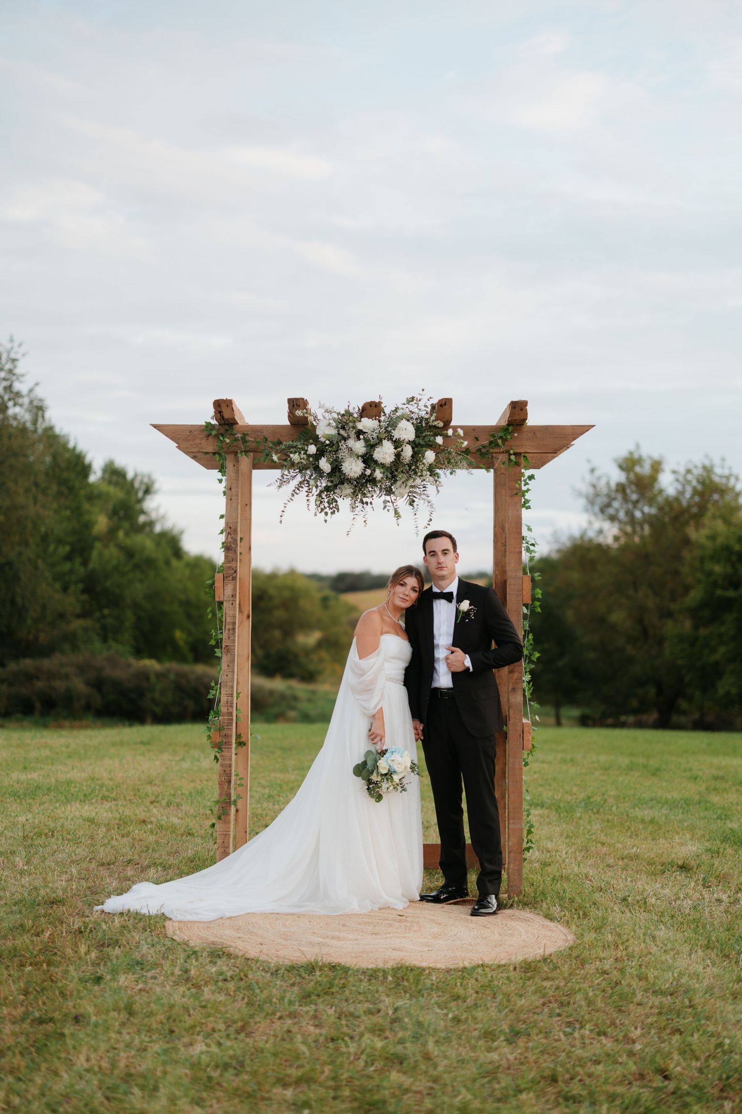 A wedding ceremony setup with wooden arch decorated with white flowers in a rustic outdoor setting.