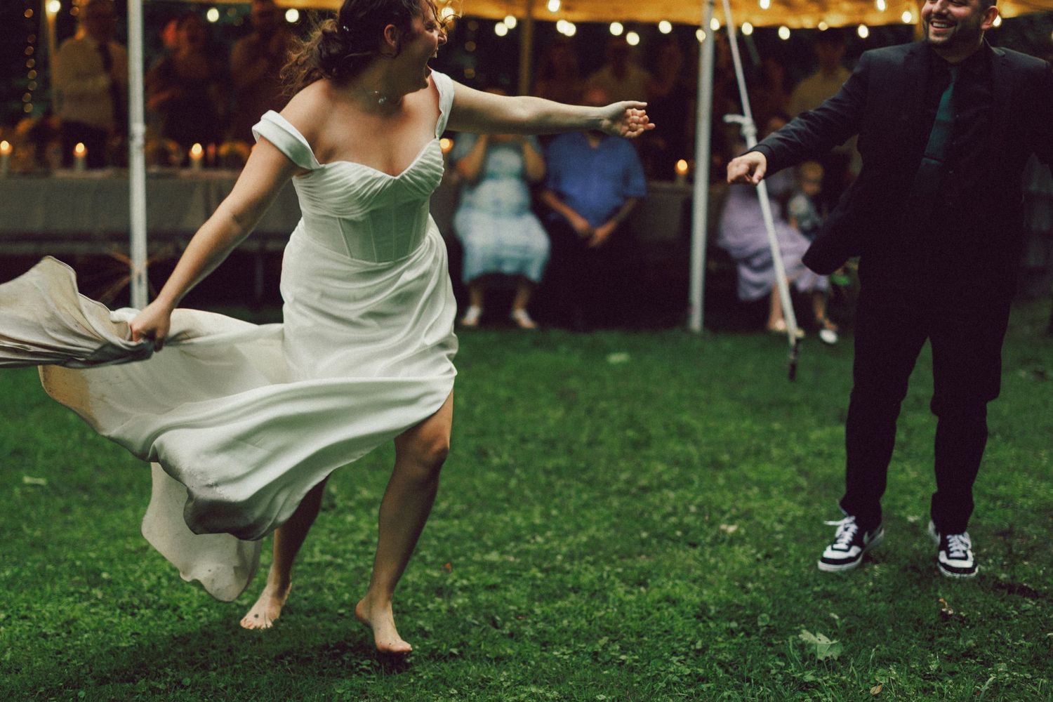 Wedding reception guests dance on grass under string lights at dusk.