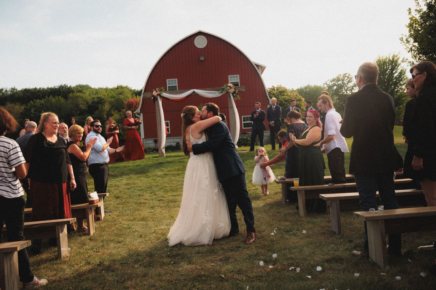 Wedding guests gather outside a rustic red barn during a sunset celebration in the countryside.