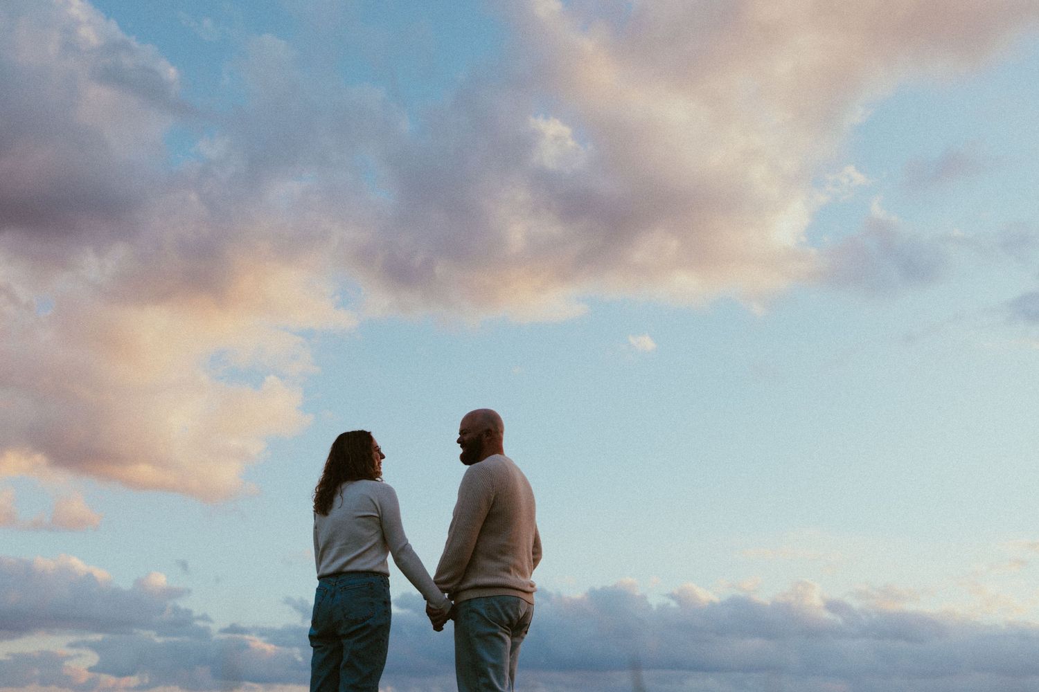 Two people stand together watching the sunset with pink clouds in the sky.