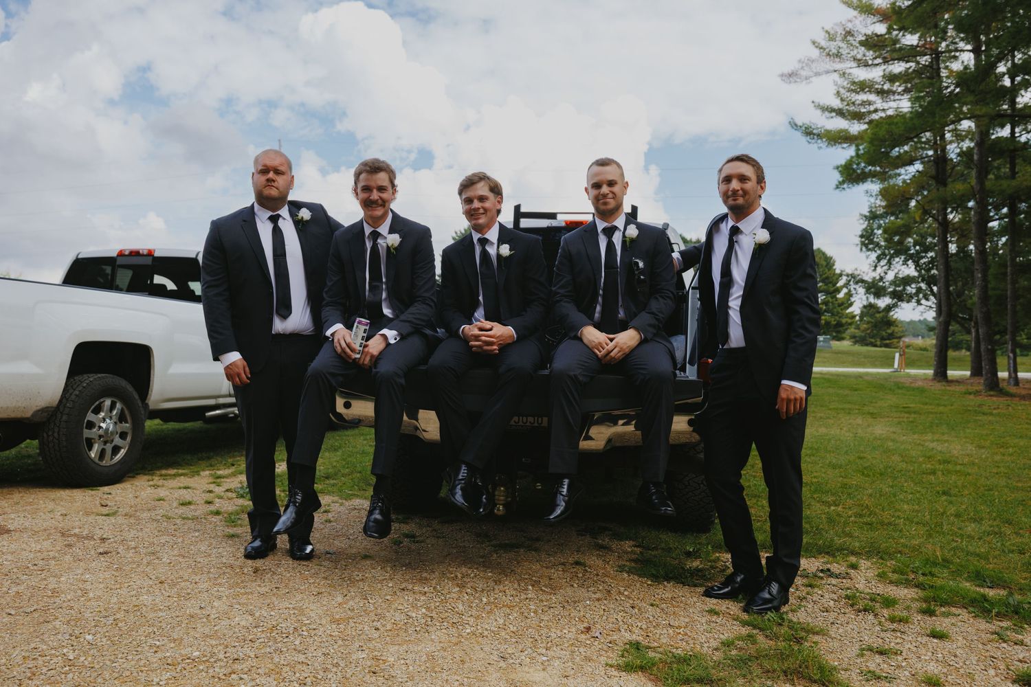 Groomsmen in black suits pose casually outdoors against a cloudy sky.
