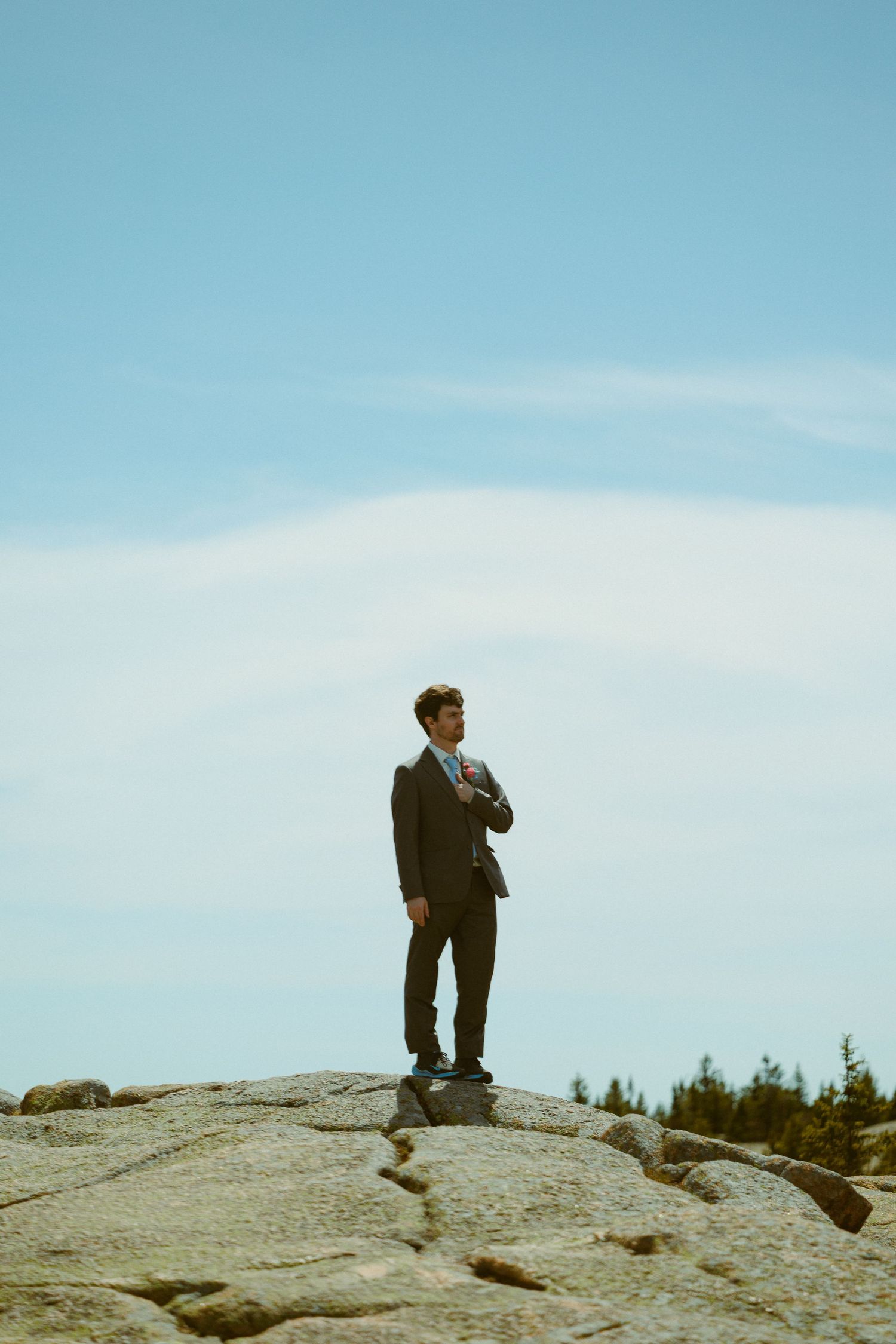 Groom in dark suit stands confidently on granite rock formation under blue sky with wispy clouds.