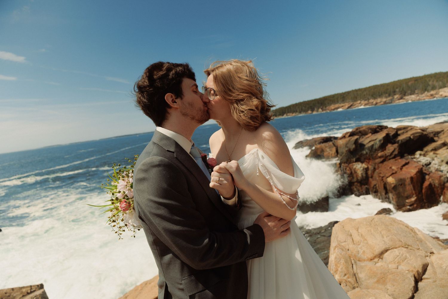 A bride and groom share a romantic kiss on rocky coastal cliffs with crashing ocean waves under a bright blue sky.