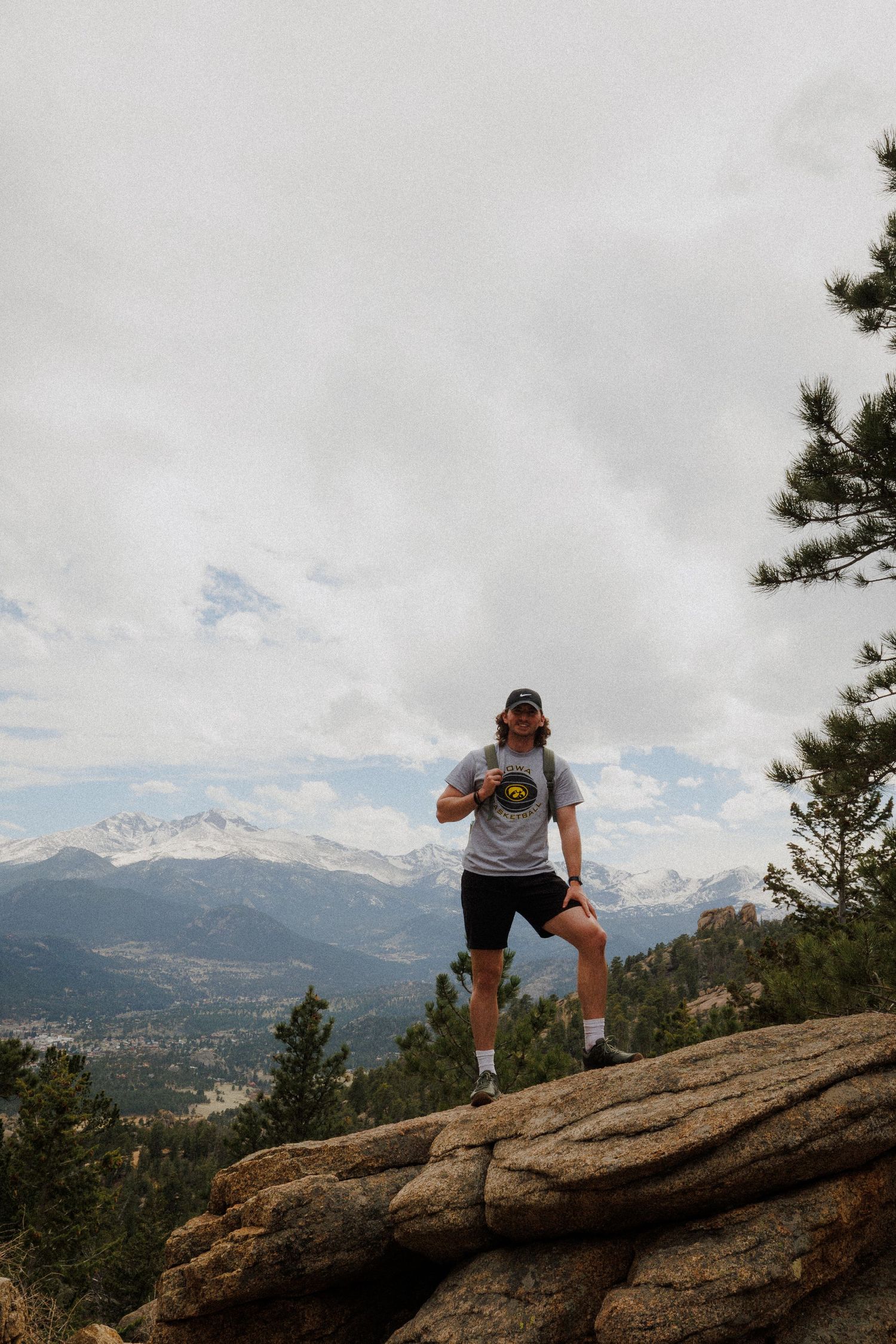Hiker climbs rocky outcrop overlooking snow-capped mountains and valley in Colorado landscape.