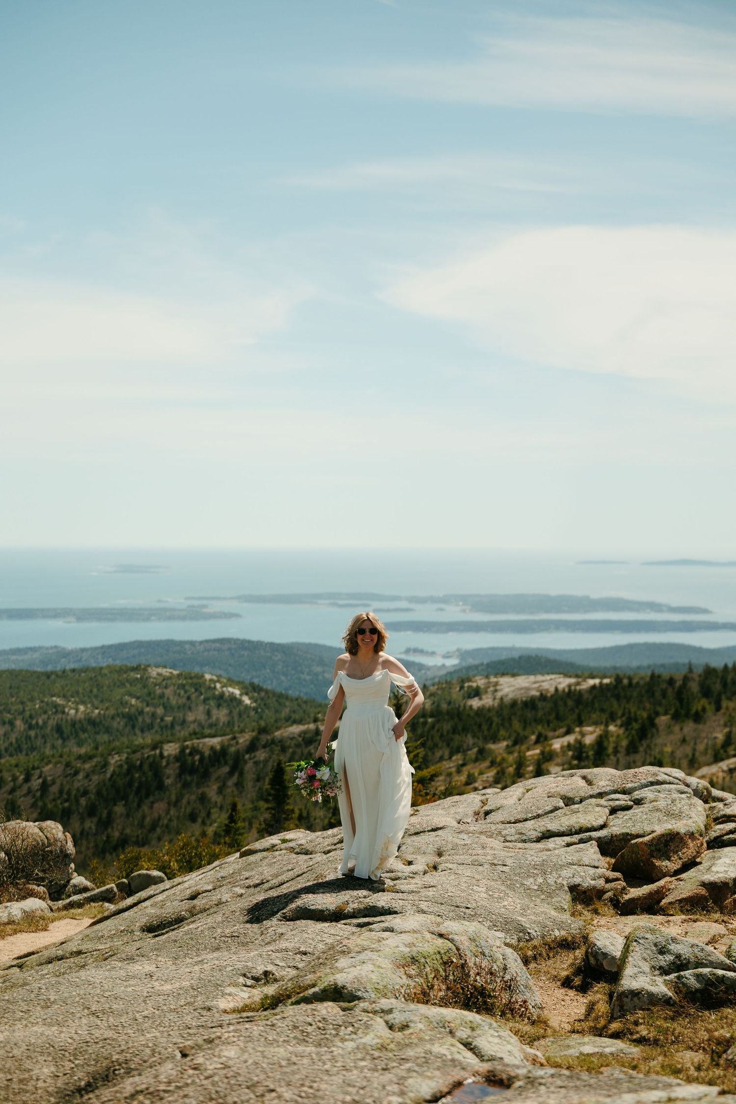 Bride in white off-shoulder gown stands on rocky mountain summit with coastal panorama.