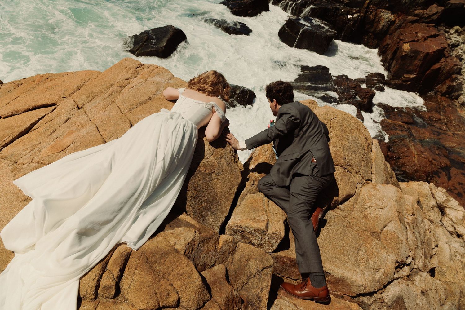 Bride and groom climbing rocky coastal cliffs in wedding attire, waves crashing below.
