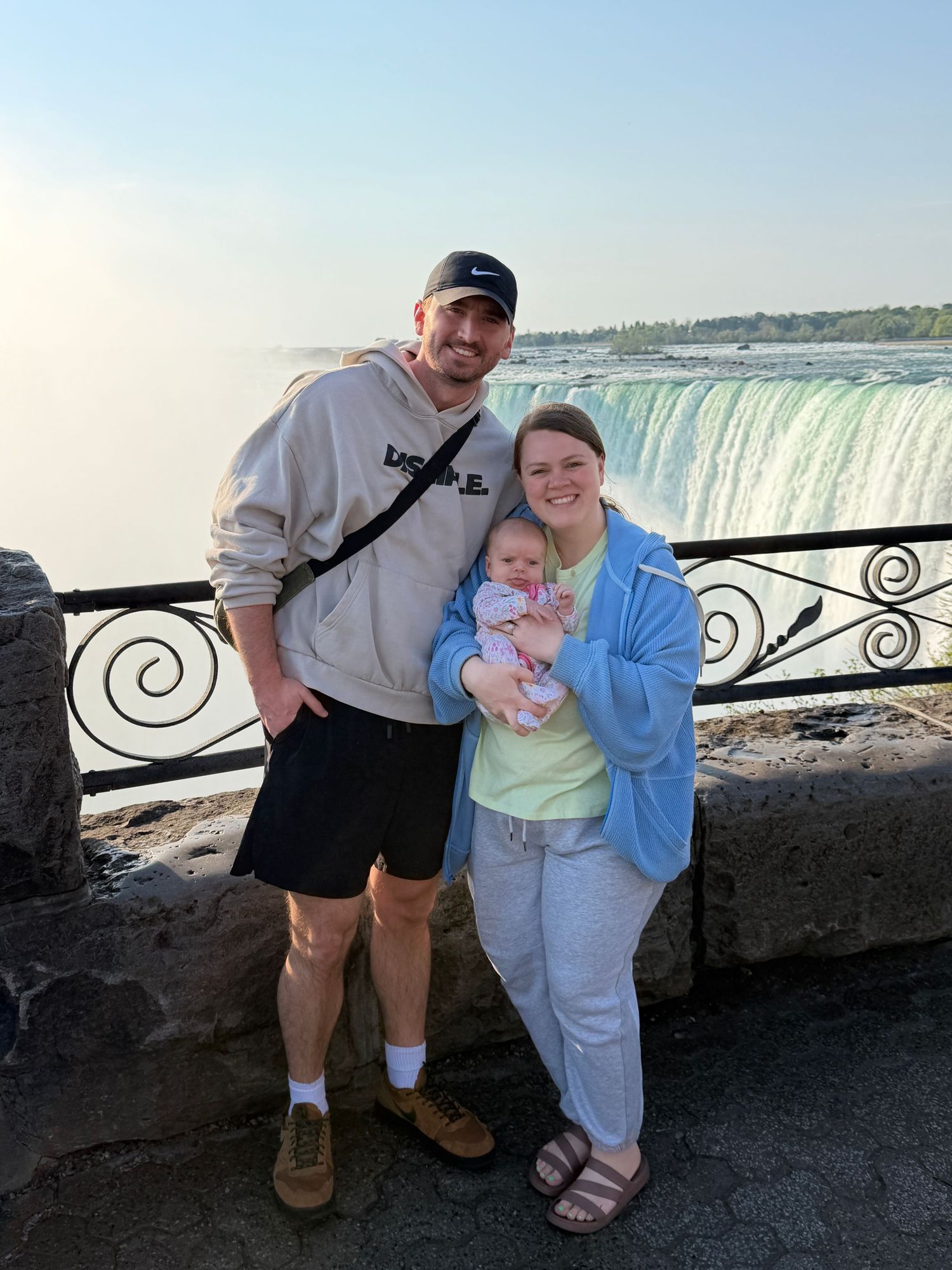 Family of three posing at Niagara Falls overlook, parents holding newborn baby with waterfall in background.