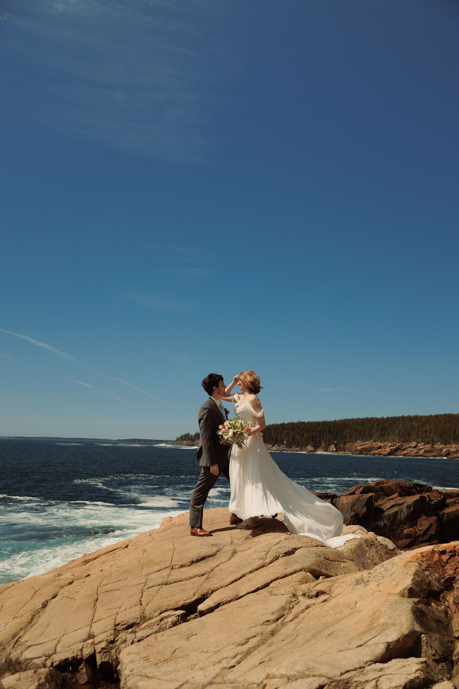 Bride and groom share a romantic moment on rocky Maine coastline with ocean waves and blue sky.