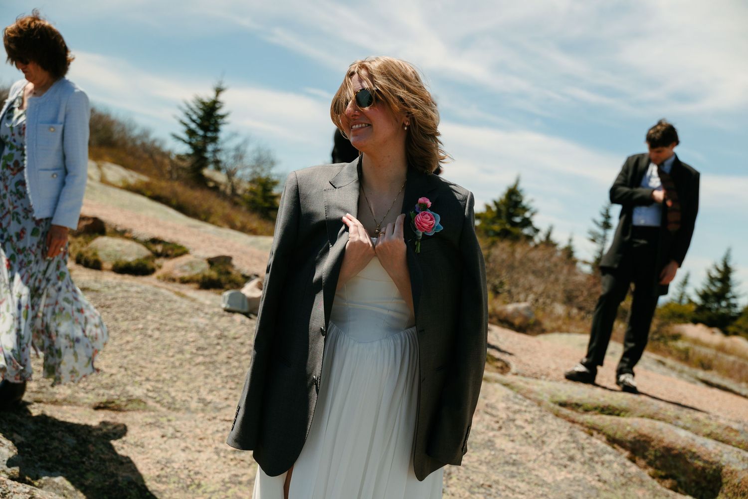 Woman in elegant gray gown and blazer with floral boutonniere smiling outdoors at a rocky wedding venue.