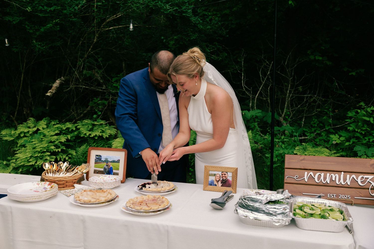 Rustic wooden dessert table at an outdoor wedding reception with multiple pies and cakes on display.